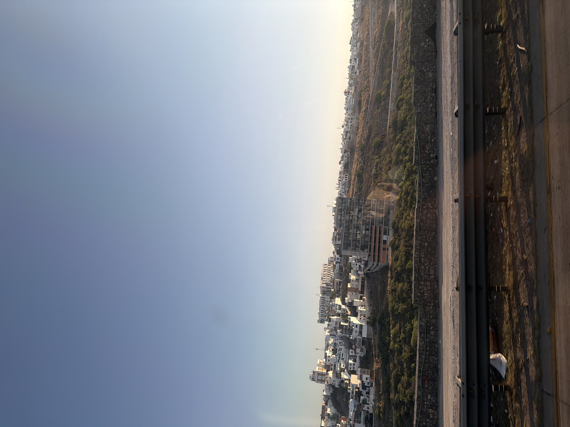 The image shows a cityscape transitioning into a hillside. In the foreground, part of a building structure with metal railings and concrete surfaces is visible, suggesting the image was taken from an elevated position. Adjacent to this is a hillside dotted with vegetation, possibly trees and shrubs, and some rocky areas. Further down the hillside, the cityscape begins, with a mix of low-rise and taller buildings. The structures appear to be mostly residential, and the buildings are clustered together. The sky above is a light blue, suggesting either early morning or late afternoon. The overall impression is that of a cityscape nestled in a hilly area, perhaps on the outskirts of a larger urban center.