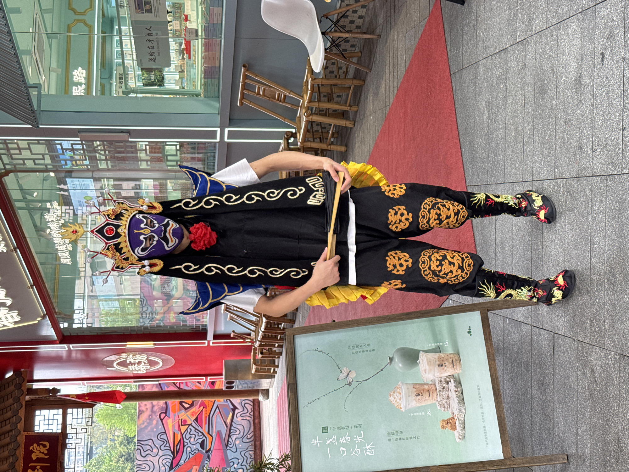 Here is a description of the image:
A person stands in a costume on a gray paved area, possibly a sidewalk or public space. The person is wearing an elaborate headpiece and mask with a red flower detail. The costume consists of a black tunic with gold detailing, black pants with similar gold accents, and black boots. The person is holding two light-colored sticks or rods.
To the person's right is a promotional display board featuring images of food and beverages, along with Chinese characters. Behind the person, there is a building with glass windows displaying a store interior. There is also red signage and what appears to be a Chinese flag. The architecture suggests that the location is in China. Based on the OCR text, there appears to be Chinese text indicating the Jinjiang district, China.