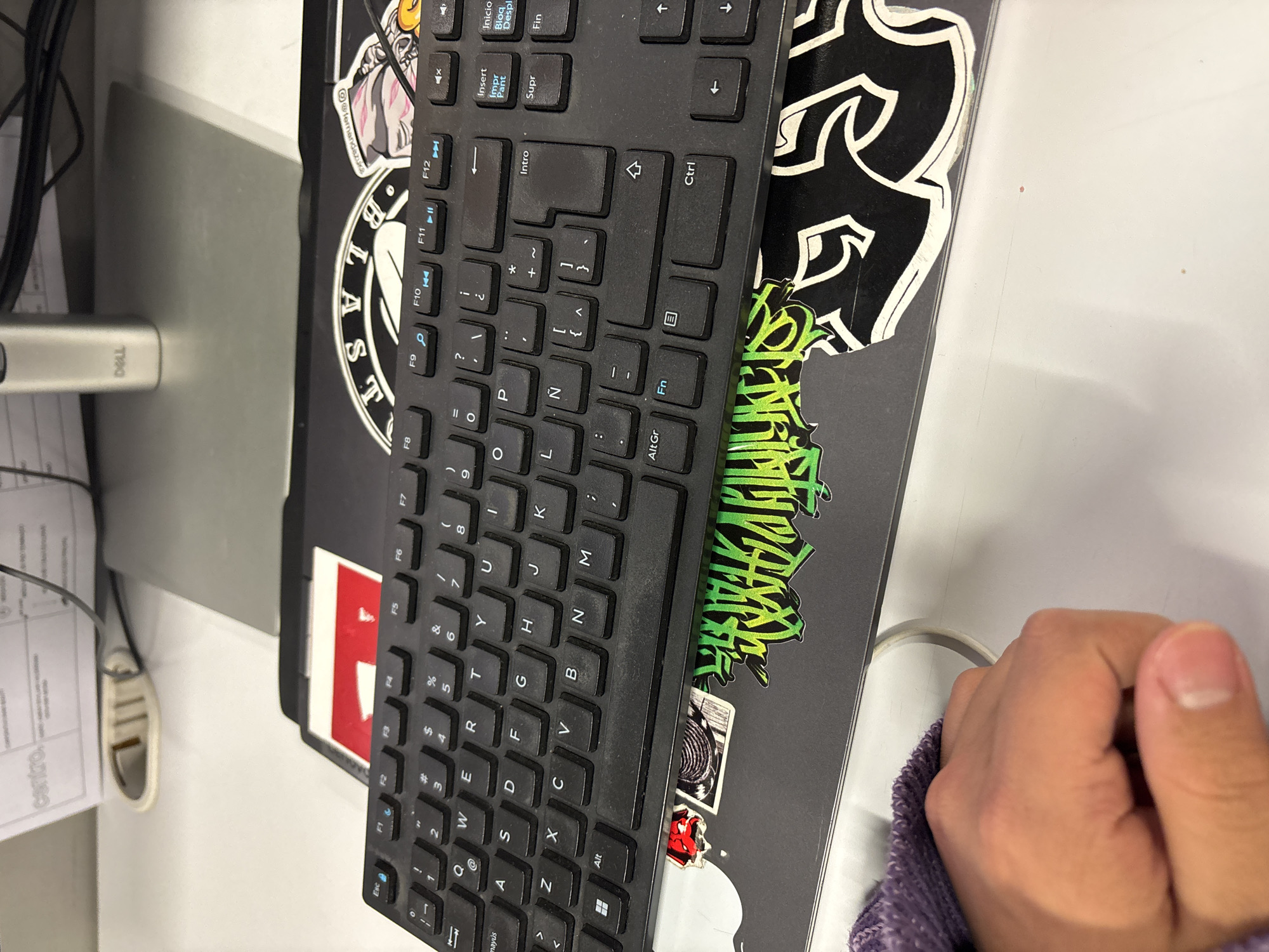 This image captures an indoor office or workspace environment in Mexico City, Mexico. The central foreground features a black computer keyboard with a Spanish QWERTY layout, showing characters such as 'Ñ' and function keys like 'Intro', 'Impr Pant', 'Inicio', 'Supr', and 'Fin'. This keyboard rests upon the lid of a dark-colored laptop, which is extensively covered with various stickers. Visible stickers include a large white and black stylized graphic, a neon green graffiti-style design, a black and white graphic depicting a figure's head, a small red sticker with a white symbol, and a cartoonish head with pink hair accompanied by the handle "@fernandovz".
In the lower-left quadrant, a light-skinned human right hand is partially visible, clutching a purple microfiber cloth. The immediate desk surface is white and appears to be made of laminate or a similar material. In the background, a silver-colored Dell monitor stand is present. Behind the monitor stand and cables, stacks of white papers are visible, one of which prominently displays the word "centro." Black electrical cables are also discernable running across the desk and connecting to unseen devices.