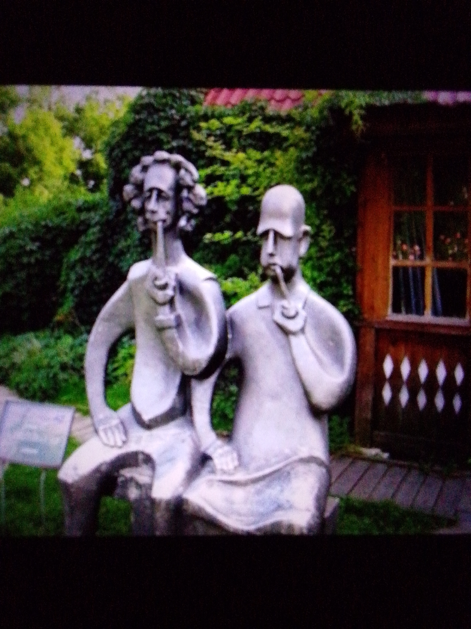Two weathered, light gray statues of men are seated on a wooden bench in a garden. The statue on the left has curly hair and is holding a pipe to his mouth. The statue on the right is wearing a cap and is also holding a pipe. Behind the statues, dense green foliage forms a backdrop, and to the right, a wooden structure with a window and decorative diamond-shaped cutouts is visible. A dark, blurry area occupies the top and bottom portions of the image.