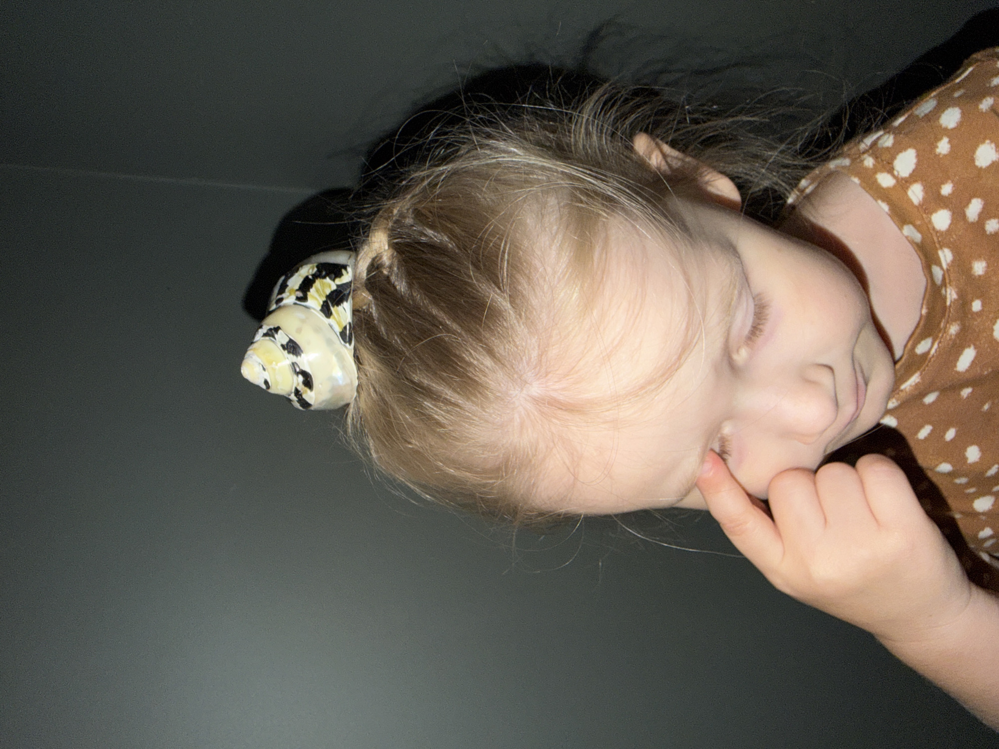 A close-up, high-angle image depicts a young child, seen from the top-right perspective, with their head tilted downwards. The child has light blonde hair, with a small braid or section pulled back on their right side (viewer's left). A hair accessory, likely a bow or clip, with a white base and a distinct black and yellow animal print or abstract pattern, is attached to this section of hair.
The child is wearing a brown garment, possibly a shirt or dress, adorned with white polka dots. Their right hand is visible, with the index finger gently touching the side of their face or ear, near the corner of their right eye. The child's eyes are open, and their gaze is directed downwards, suggesting a state of contemplation or curiosity, perhaps looking at something off-camera.
The background is a plain, dark gray or black surface, providing a simple and unobtrusive setting. The lighting is soft, highlighting the child's features. The overall scene conveys a quiet moment. There is no visible text in the image. The original image is rotated 90 degrees clockwise.