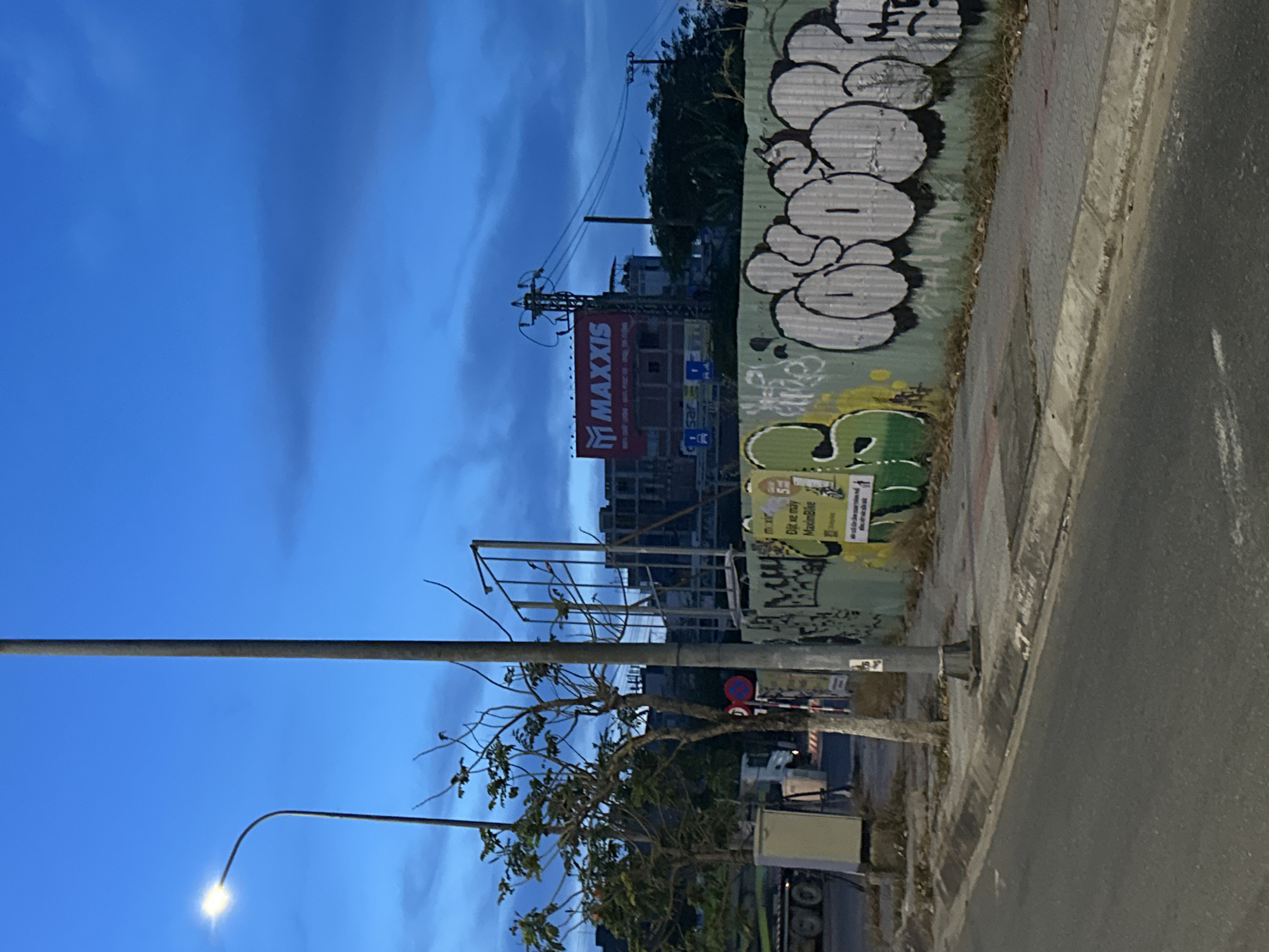 The image depicts a street scene at dusk in Da Nang, Vietnam. A street lamp is illuminated on the left side of the frame, casting light onto a sidewalk. A tall, thin pole stands in the center, likely another street light or utility pole. Behind the pole, a construction site is visible with scaffolding and partially built structures. A red billboard with the "MAXXIS" logo is prominently displayed on one of the buildings. The wall of the construction site is covered in graffiti, featuring stylized lettering and artwork in shades of green, white, and black. A small tree with bare branches grows on the left side of the sidewalk, near a beige utility box. A traffic sign with a red circle is mounted on a pole. The road in the foreground is paved and appears to be a city street. The sky is a deep blue with scattered clouds, indicating the transition from day to night.