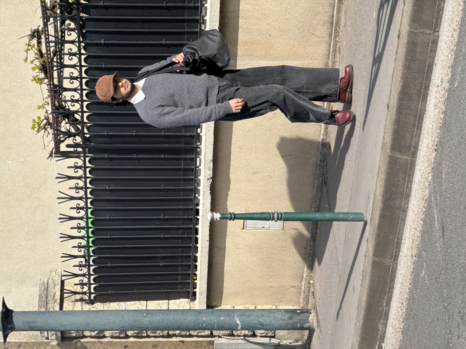The image depicts a lone adult male walking on a concrete sidewalk in Asnières-sur-Seine, France. The individual, centrally positioned and facing right, is captured in motion. He is dressed in a brown corduroy baseball cap, dark sunglasses, a white collared shirt beneath a gray crew-neck sweater, dark gray wide-leg jeans, and dark red lace-up boots. A black duffel bag is carried over his left shoulder. To the left, a green ornamental bollard with a polished cap stands on the sidewalk, casting a long shadow across the pavement. A dark green utility pole is partially visible on the extreme left. The background features a continuous light beige stucco wall, surmounted by a black wrought iron fence with decorative spiked finials. Above the fence, minimal green foliage is present. A small, light-colored electrical junction box is affixed to the stucco wall behind the bollard. The immediate foreground includes a dark asphalt road bordered by a concrete curb. The scene is brightly lit by direct sunlight, producing sharp, elongated shadows on the ground.