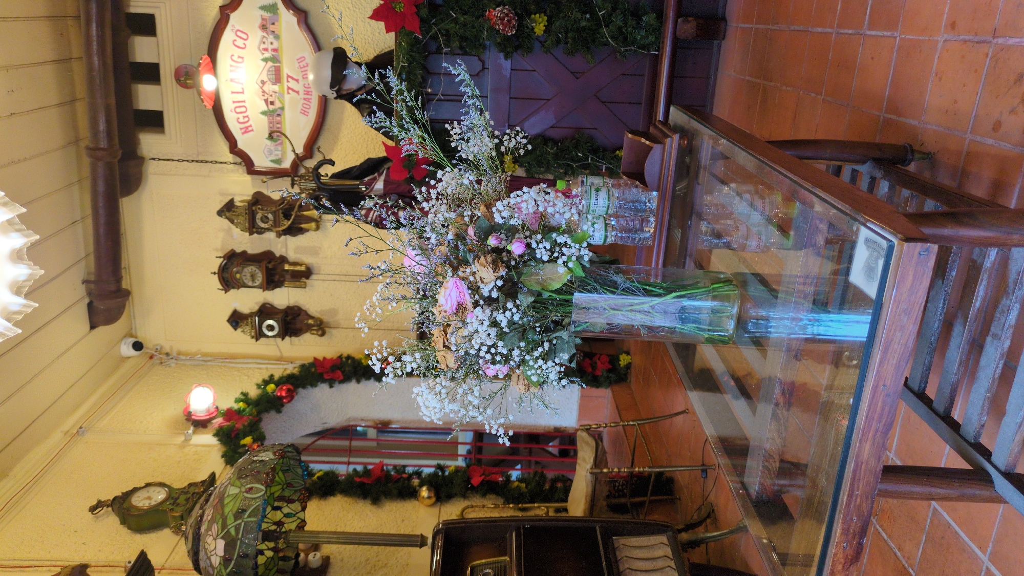 This is a medium shot of the interior of a cafe in Da Lat, Vietnam, featuring a glass-topped wooden coffee table in the foreground. On the table is a clear glass vase filled with a large bouquet of dried and fresh flowers, predominantly white baby's breath and some pink and brown roses. Two plastic water bottles are also on the table. The background reveals a cluttered, vintage-themed space. To the left, a stained-glass lamp with a floral design sits on a wooden stand. Further back, a Christmas garland with red bows and baubles is draped over an arched doorway. Several antique wall clocks are visible on the yellow wall, along with a sign that reads "NGÔI LÀNG CỔ 77 HOÀNG DIỆU". A person wearing a white helmet and a mask is partially visible on the right, near a wooden door decorated with a Christmas garland and poinsettias. The floor is tiled with terracotta-colored squares. The lighting is warm and ambient, suggesting an indoor setting.