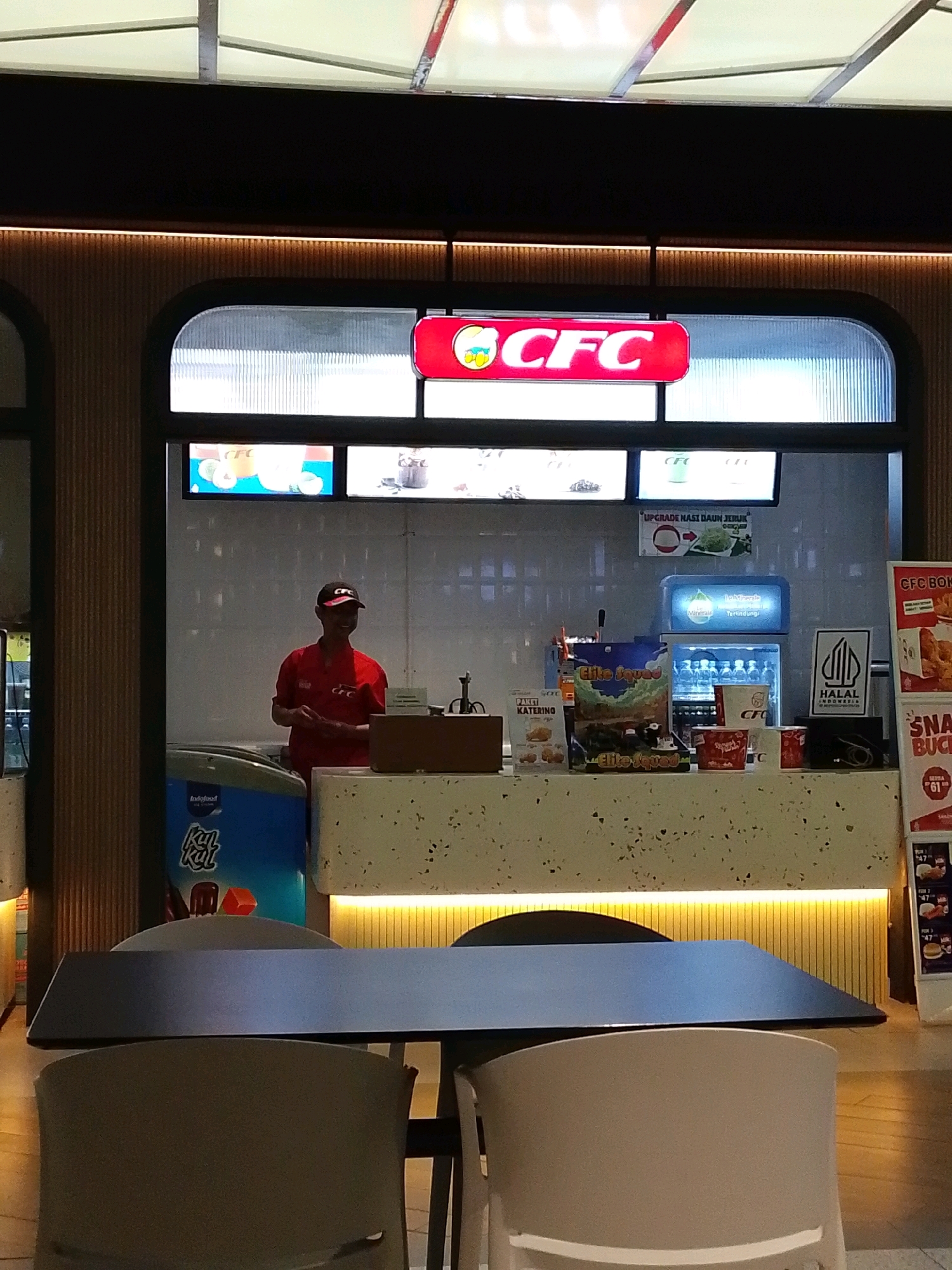 This image shows the interior of a fast-food restaurant, likely located in Semarang, Indonesia, based on the product labeling and language visible. The scene features a counter with various food and drink items displayed. A man in a red uniform and a baseball cap with a "CFC" logo is standing behind the counter, appearing to be an employee. In the foreground, there are tables and chairs, suggesting a dining area. The overall lighting is bright, with a mix of overhead and accent lighting creating a well-lit environment. The restaurant appears to be clean and modern, with a focus on displaying menu items and branding.