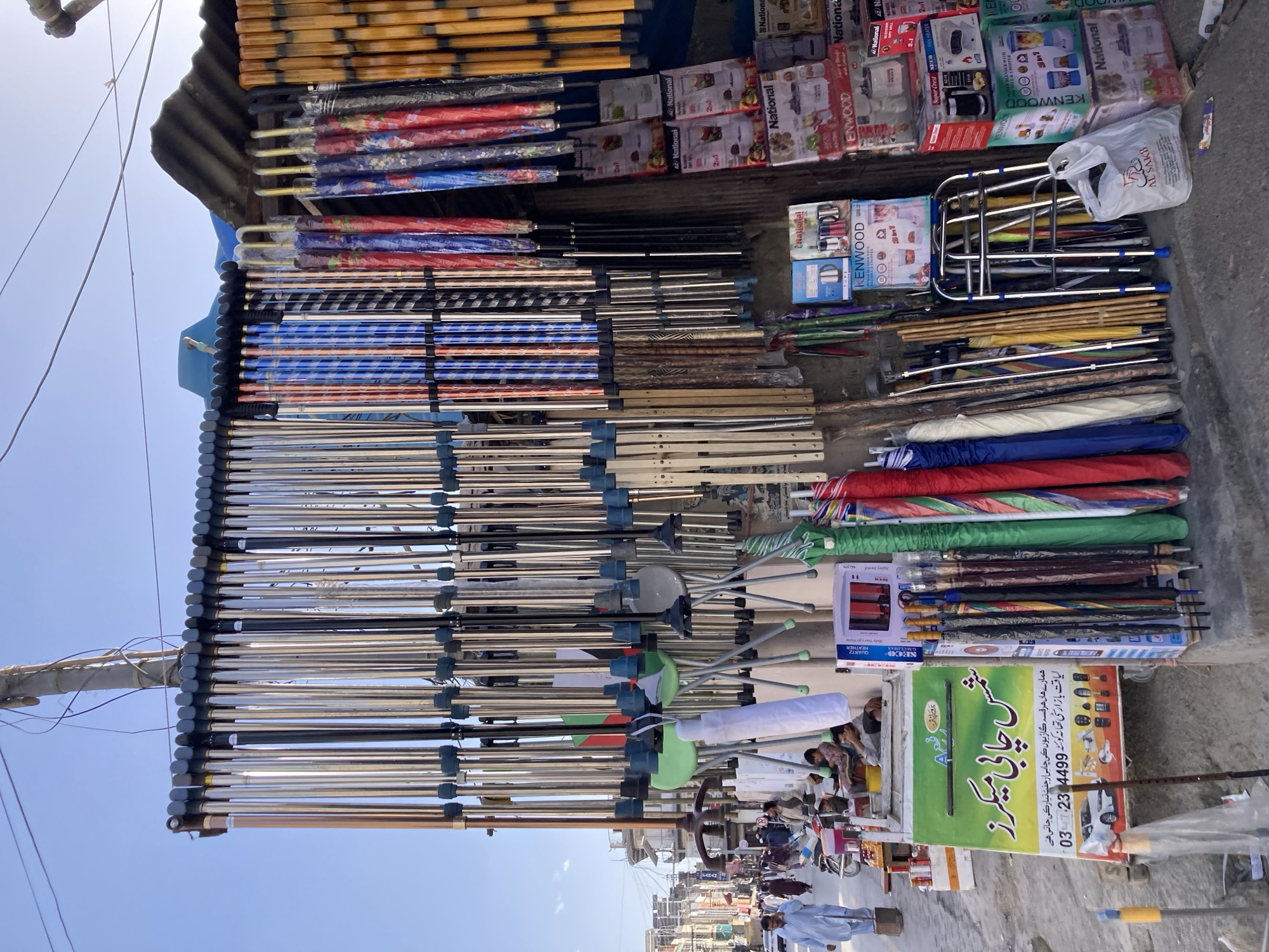 The image captures a street scene in Quetta City Tehsil, Pakistan. A stall is displaying a variety of items, including umbrellas stacked in rows, kitchen appliances in boxes (marked with brands like "National" and "KENWOOD"), and what appears to be furniture. A sign for "Shams Chabi Makers" (Shams Key Makers) is visible, advertising key-making services for cars. People are visible in the background, suggesting an active street environment.