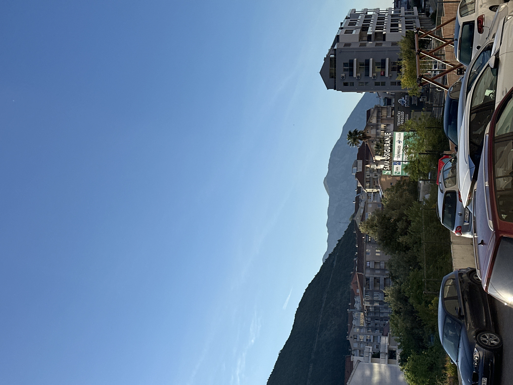 The image shows a view in Budva, Montenegro. A clear, blue sky takes up the majority of the left side. In the right half, there is a mountain range visible in the background. In the foreground, there are several parked cars partially visible, lining a street. Behind the cars, a mix of buildings including residential apartments, commercial signs (like "Sava Osiguranje"), and trees suggest an urban environment. A grey modern-style building can be seen further back on the right.