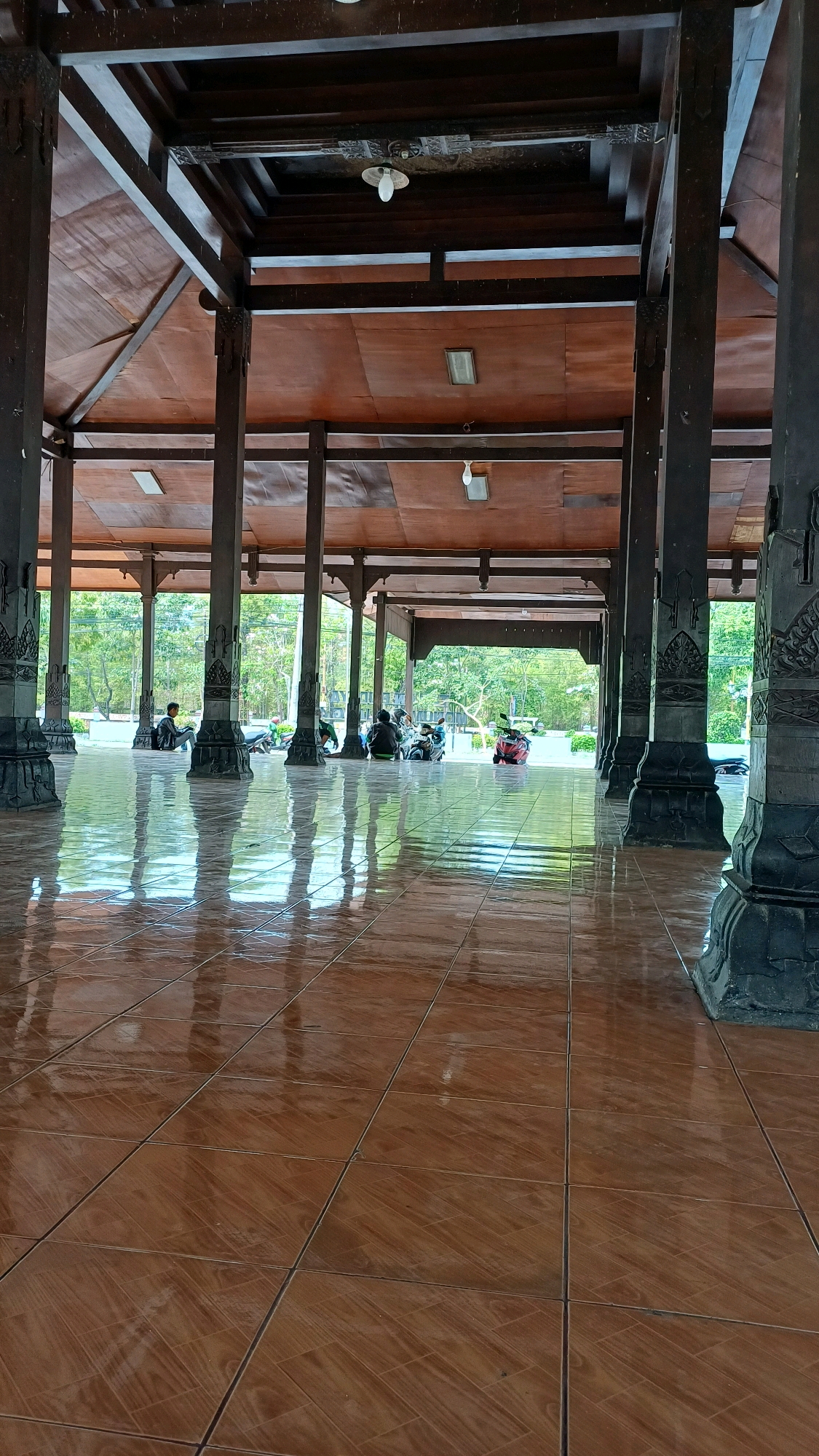 The image shows a wide, open-air pavilion with a brown tiled floor that is highly reflective, showing the dark wooden pillars and ceiling beams, as well as the green trees and sky outside. Several people are seated or resting near the edge of the pavilion, with motorcycles parked behind them. The pavilion appears to be in a natural, outdoor setting, possibly a park or recreational area, located in Bareng Lor, Indonesia. The lighting suggests it is daytime, with natural light filtering in from the open sides. The overall atmosphere is peaceful and relaxed.