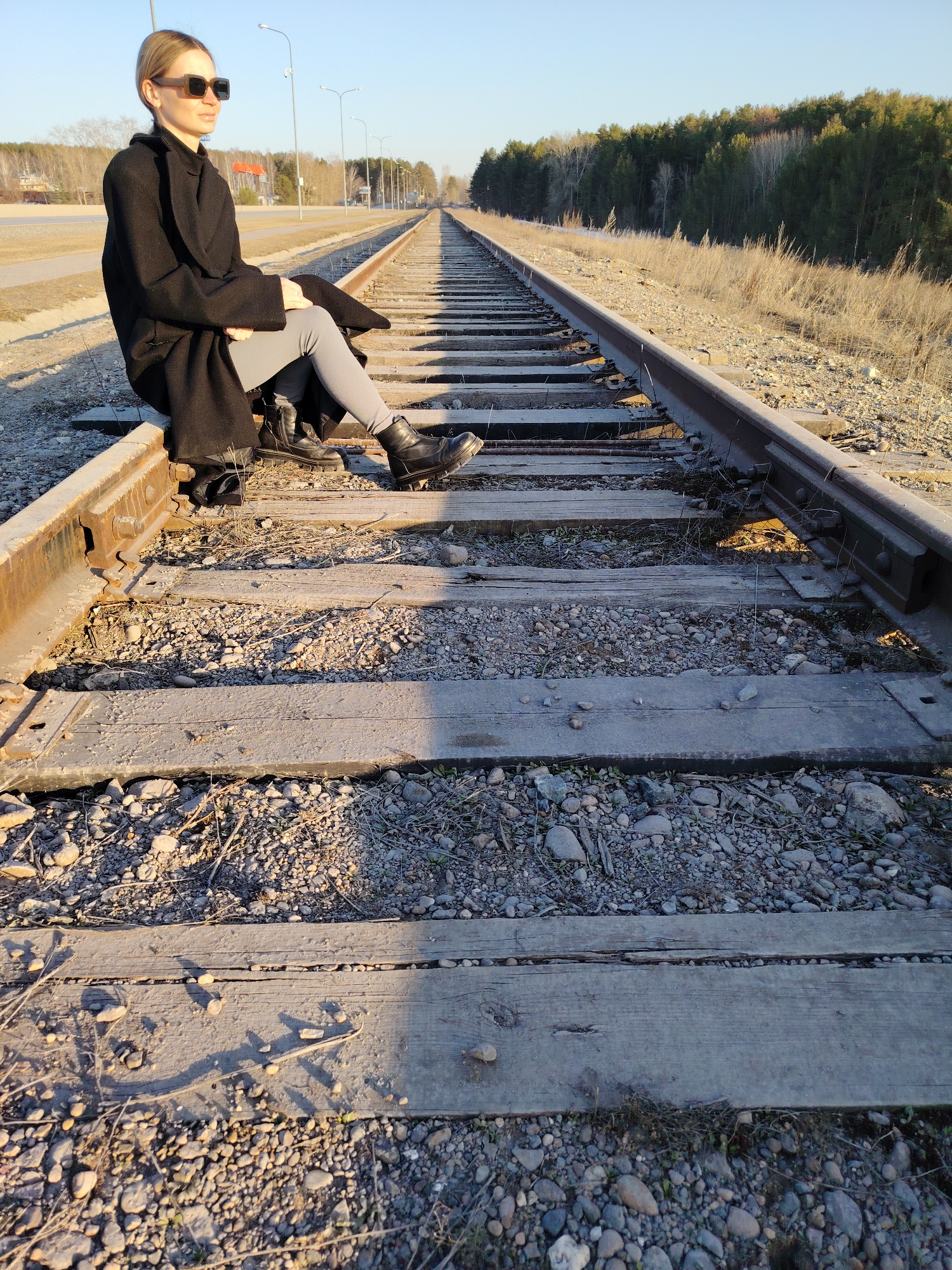 A woman with blond hair and wearing sunglasses sits on the left rail of a railroad track. She is wearing a black coat, gray leggings, and black boots. The railroad track extends into the distance, with trees on the right side of the track. The location is Chaikovsky, Russia.