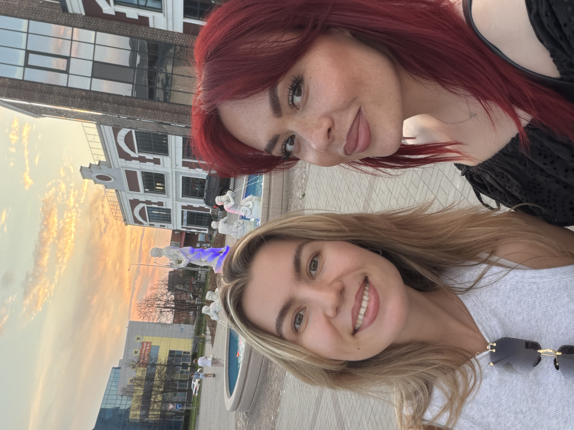 Two women are taking a selfie outdoors at dusk in Tolyatti, Russia. The woman on the left has blonde hair and is wearing a grey top with sunglasses hanging from her neckline. The woman on the right has vibrant red hair and is wearing a black off-the-shoulder top. They are standing in front of a fountain with sculptures and a background of modern buildings with brick and glass facades, characteristic of Tolyatti's newer architectural developments. The sky is painted with warm hues of orange and yellow from the setting sun. In the middle ground, there are people walking and a white car. The ground is paved with light-colored square tiles. The overall scene suggests a pleasant evening in an urban setting.