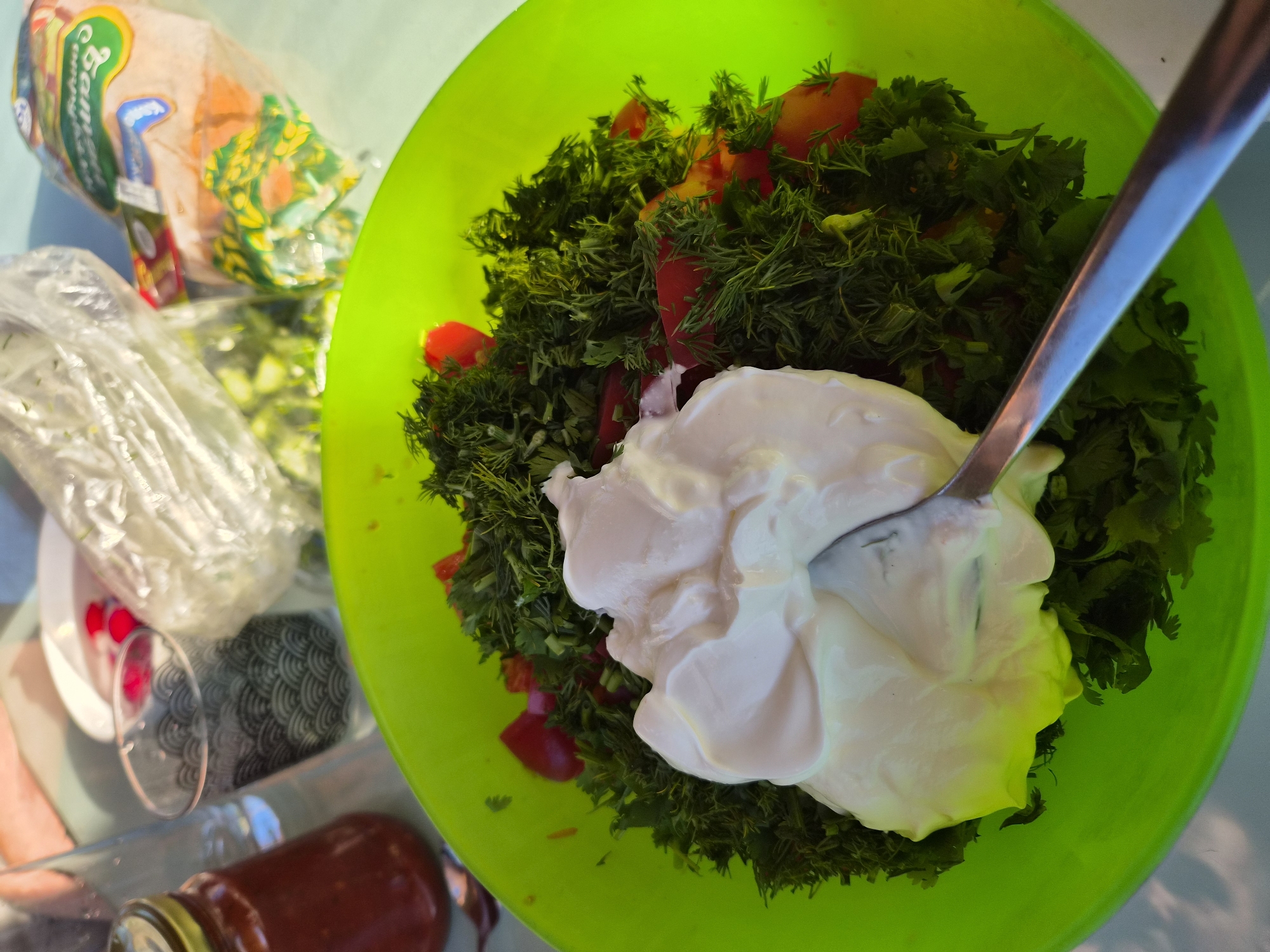 The image displays a high-angle view of food preparation taking place in Motyakovo, Russia. In the foreground, a bright lime-green plastic bowl contains a salad composed of finely chopped green herbs, including dill and cilantro, mixed with diced red tomatoes. A large dollop of white, creamy dressing is situated centrally in the salad, with a silver spoon partially submerged and positioned diagonally, indicating that the dressing is being stirred into the ingredients.
In the background, to the left, a reddish-brown jar with a gold lid is visible next to a clear, empty drinking glass. A person's hand and forearm are partially visible at the upper left. Towards the center, a clear glass with a dark, wave-pattern design stands alongside a small white bowl containing several small, round red items. Further back, two plastic bags are present: one appears open and contains chopped green vegetables, while the other is a sealed package of bread, clearly labeled with Cyrillic text including "Батон" (Baton), and features green and yellow packaging elements. The entire scene is set on a light blue or green surface, illuminated by bright natural light.