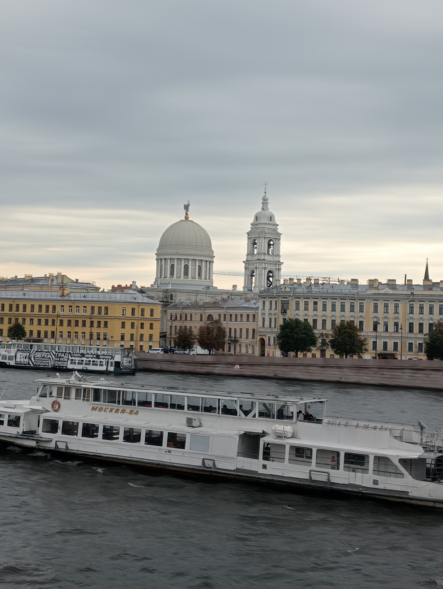 This is a vertical, medium-shot photograph of a riverfront cityscape in Saint Petersburg, Russia, under an overcast sky. In the foreground, a large, white passenger boat with multiple decks and large windows is traveling on the dark, choppy water. The boat has the Cyrillic lettering "МОСКВА-84" on its side. Behind the boat, a river bank is visible, lined with trees and buildings.
The architecture in the background includes a prominent large, light-colored dome with a golden statue on top, and a tall, white bell tower with arched openings. These structures are part of a larger complex of pale yellow and cream-colored buildings with numerous windows and ornate details. The overall impression is of a historic and grand urban setting. The lighting is diffused due to the cloudy sky, creating a somewhat muted color palette.