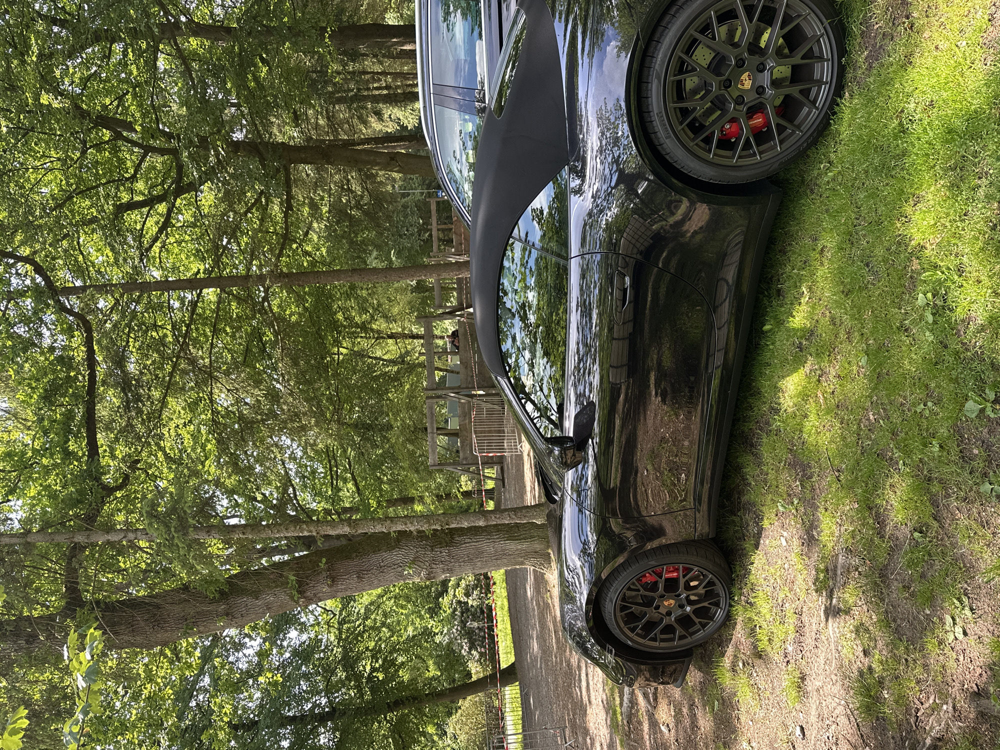 The image depicts a black Porsche 911 convertible, likely a 992 generation, parked on a grassy verge adjacent to a dirt path in a densely wooded recreational area. The car's black soft top is raised. It features dark multi-spoke wheels with visible red brake calipers, indicating high-performance brakes. Pirelli tires are mounted on the wheels. Sunlight filters through the overhead tree canopy, casting dappled light and shadows across the scene and reflecting off the car's glossy bodywork.
In the mid-ground, a dirt or gravel path is visible, bordered by a temporary barrier consisting of metal posts and red-and-white striped tape. Beyond this barrier, towards the left, are more trees and green foliage, including what appears to be rhododendron or similar flowering shrubs. Towards the right background, behind the Porsche, a dark-colored SUV or wagon is partially visible, parked. Further back in the center-right, large, rustic wooden structures, consistent with a playground or an adventure course, are present. A faint human figure is discernible sitting on a distant wooden bench or structure behind these wooden elements. The ground consists of patches of green grass interspersed with exposed soil and leaf litter.
The scene suggests a **wooded recreational area or a 'Waldspielplatz' (forest playground) within the city limits of Oberndorf am Neckar, Germany**.