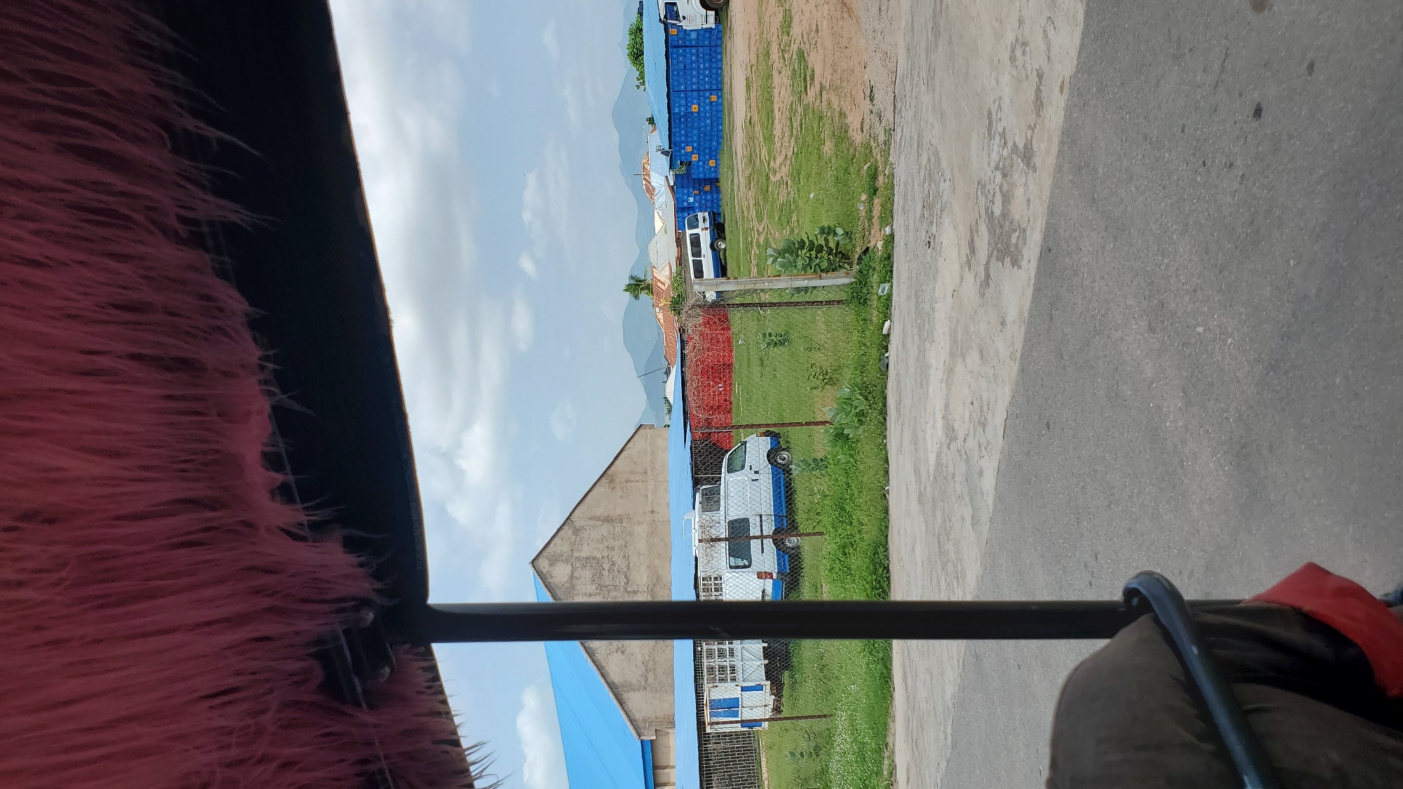 This media file depicts an outdoor scene in Jalingo, Nigeria, likely during the daytime under a partly cloudy sky. The perspective is from within a vehicle, with a furry, pink-colored material obscuring the lower left portion of the frame, possibly a decorative covering inside the vehicle. A dark metal bar runs horizontally across the lower center of the image, suggesting the viewer is looking over or through a part of the vehicle's interior.
The background reveals a somewhat industrial or storage area. Stacked shipping containers, predominantly blue and red, are visible in the middle ground, suggesting a logistics or distribution hub. Several white vans or small trucks are parked amongst the containers, some of which are partially obscured. A chain-link fence separates the immediate foreground from the area with the containers and vehicles. In the further distance, mountains can be seen under the sky, with some sparse vegetation and buildings. The ground in the foreground appears to be a mixture of concrete and asphalt.
No specific activity or event is clearly taking place, with the scene appearing to be a static view of the surroundings. There is no visible text within the image. The overall impression is one of a functional, somewhat utilitarian environment, potentially a port, warehouse, or similar facility.