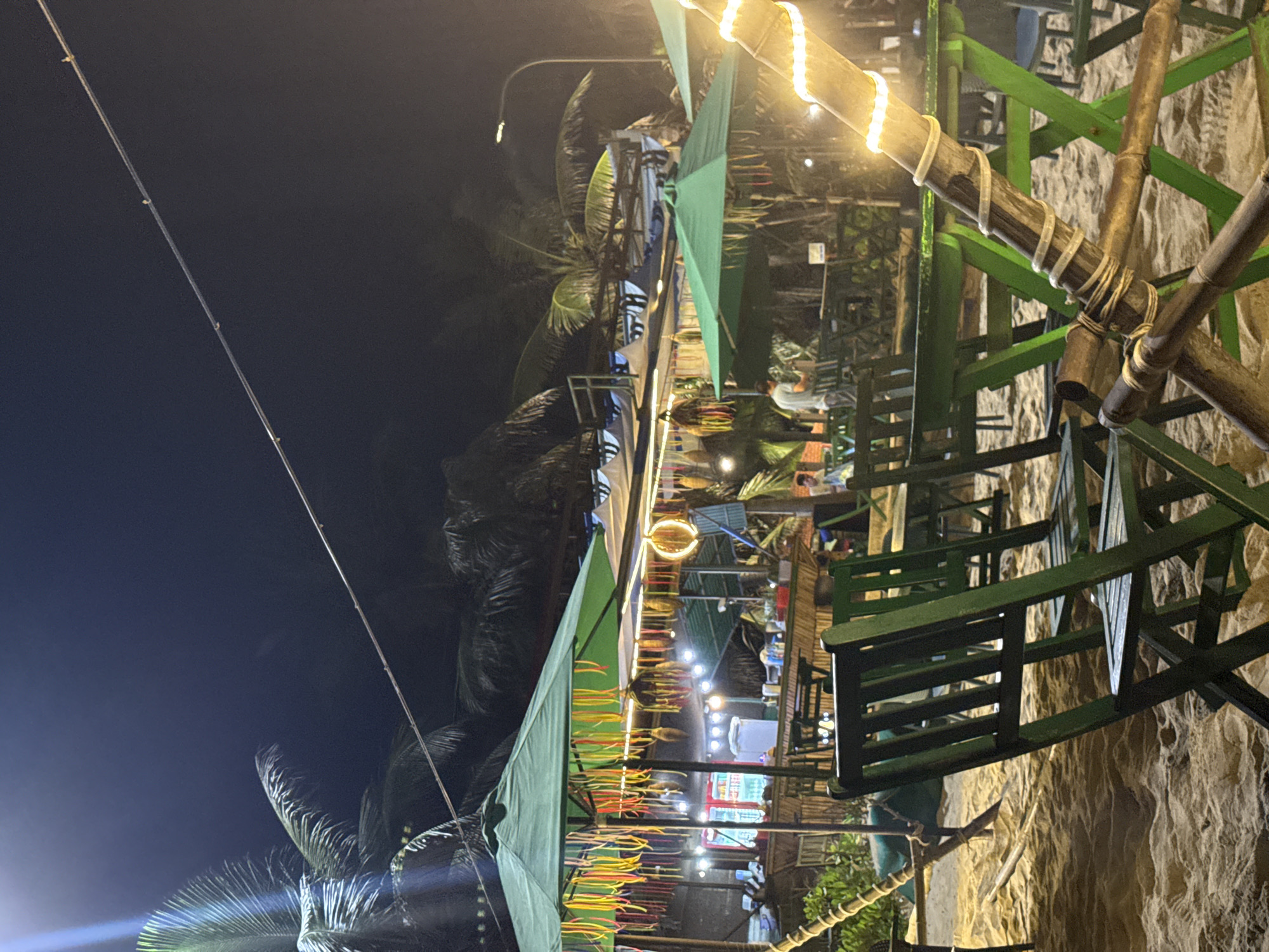 At night, a sandy area in Đà Nẵng, Vietnam is furnished with green, bamboo furniture and decorative lights. The sandy ground indicates proximity to a beach. The bamboo furniture is arranged in rows and columns, suggesting an outdoor seating area, possibly for a bar or restaurant. Strings of lights are wrapped around the bamboo poles, and additional lights can be seen further back, illuminating what looks like a building facade or interior. Palm trees are visible in the background. An electrical wire runs across the sky. The overall environment is dimly lit with artificial light sources.