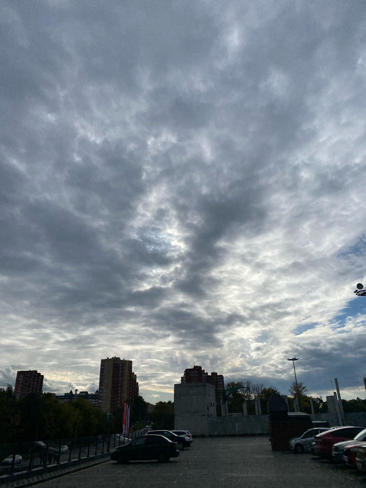 The image depicts a grey, overcast sky filled with layered clouds, with shafts of sunlight breaking through in places, creating a dramatic and atmospheric effect. Below the sky, a city skyline is visible, characterized by residential buildings with reddish-brown facades. Some of these buildings are tall and appear to be apartment complexes. In the foreground, there is a paved parking lot with several cars parked. The ground is a dark, textured surface, likely asphalt or paving stones. To the left and behind the parking lot, there is a line of trees, some with autumn foliage, and a road or walkway bordered by a dark railing. In the background, the construction of what appears to be a new building or structure is evident, with concrete columns and walls. The scene suggests an urban environment in Ivanteyevka, Russia, under a moody sky. There are no people clearly visible in the image.