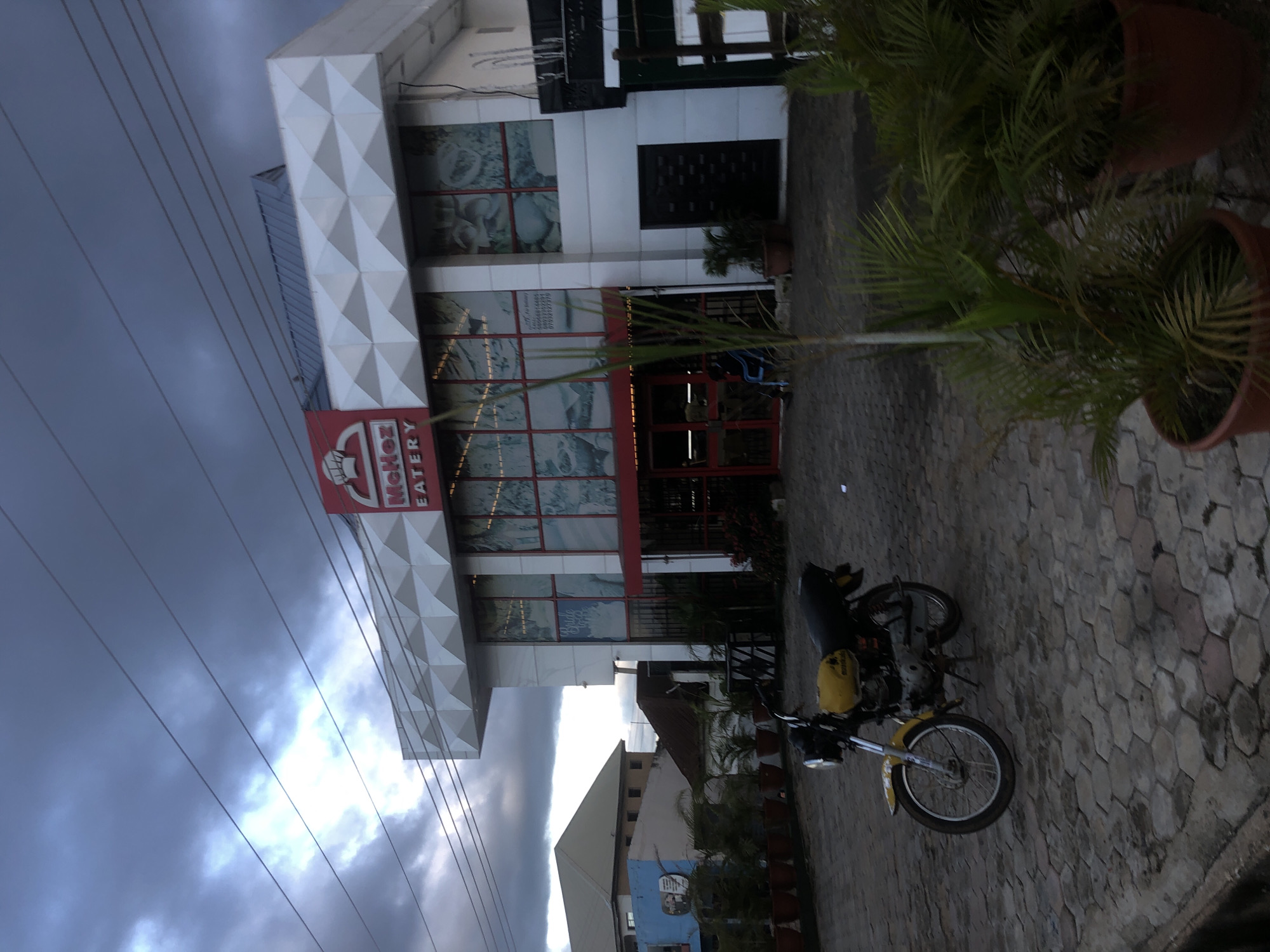 The image shows a modern, multi-story eatery building with a white, geometric facade and large glass windows. A red sign with "MCKez EATERY" is visible at the top. The building is situated in what appears to be a commercial area in Barakin Lerawi, Nigeria, with a paved courtyard in front. A yellow and black motorcycle is parked on the right side of the foreground. Several potted plants are placed around the entrance of the eatery and to the right of the motorcycle. In the background, other buildings and a cloudy sky are visible. The overall lighting suggests it is late afternoon or early evening.