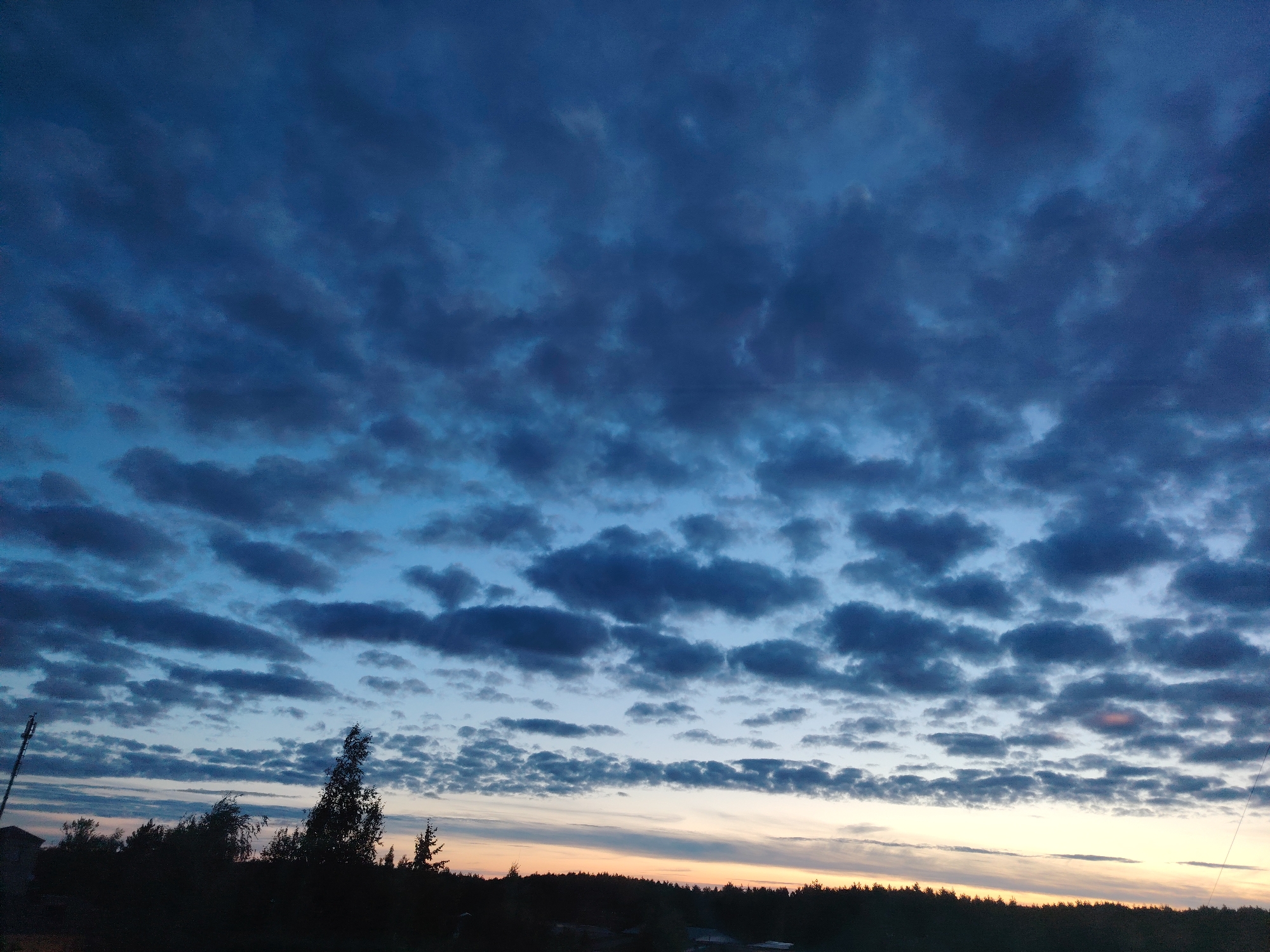 A twilight sky with scattered dark clouds is visible over a silhouetted tree line and a few structures. The sky transitions from a deep blue at the top to lighter shades of blue and hints of orange near the horizon. The clouds are a mix of larger, darker formations and smaller, lighter ones, creating a textured pattern across the sky. The lower portion of the image shows a dark silhouette of trees, some appearing windblown, along with the faint outlines of buildings and what appears to be a tall antenna or pole on the left side. The overall impression is of a serene, natural landscape at dusk.