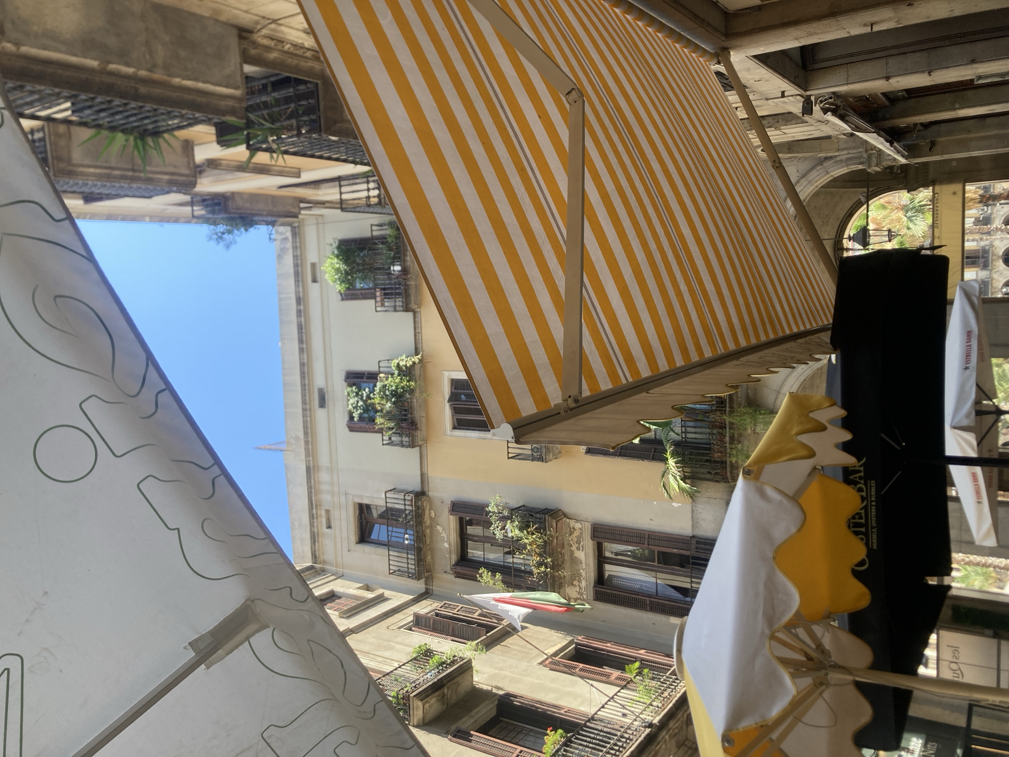 This image shows a street scene in Barcelona, Spain, likely in an older part of the city with narrow streets and historic buildings. The perspective is looking upwards, as if the photographer is standing at street level.
In the foreground, there are several awnings and umbrellas providing shade. A white and yellow umbrella with a scalloped edge is prominent on the left. Behind it, a black umbrella with "OYSTER BAR" and "MUSSELS, OYSTERS & BUBBLES" printed on it is visible. A yellow and white striped awning extends across the top right of the frame, supported by a metal arm.
The background features the facades of several multi-story buildings. The building on the left is a weathered stone color, with multiple windows and balconies adorned with plants. A flag with vertical stripes of green, white, and red is hanging from one of the balconies. The building in the center is lighter in color, with balconies also featuring greenery. The building on the right is also made of stone and has balconies. The architecture suggests a European city, and the presence of a flag with these colors points to the possibility of a region or country within Europe. The overall impression is a charming, sunny day in a Mediterranean city.
The image is well-lit, indicating it was taken during daylight hours, likely with a clear sky, as suggested by the bright blue visible between the buildings. No people are explicitly visible in the frame, but the presence of outdoor seating and awnings implies a commercial area, possibly a cafe or restaurant.