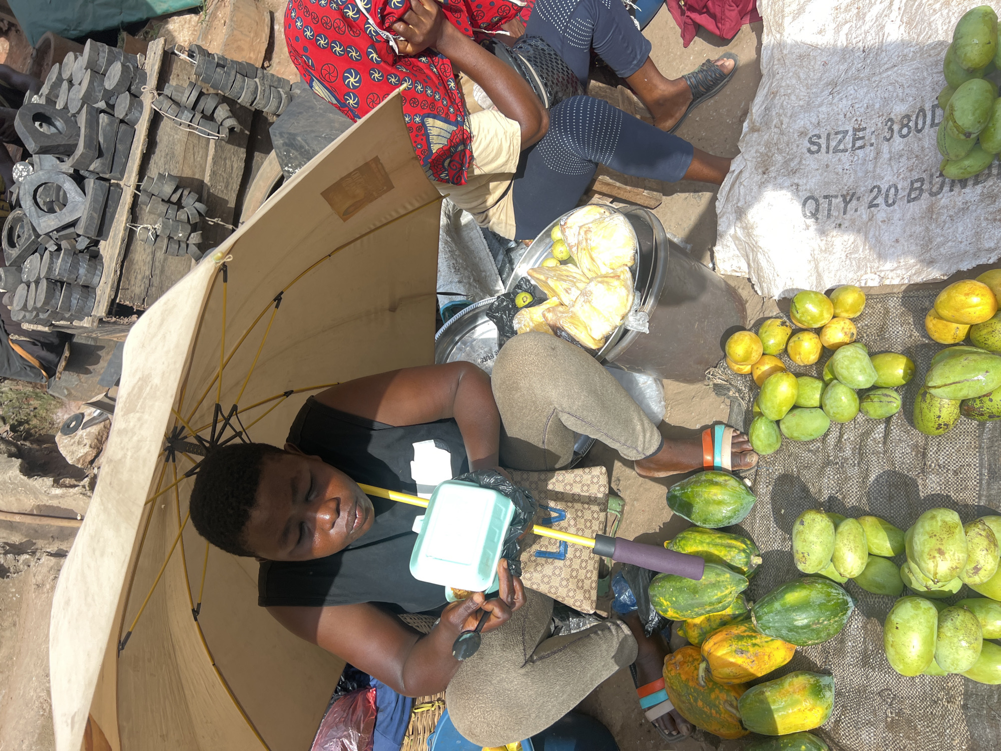 This media file captures an active outdoor market scene in Nkpor, Nigeria, under bright daylight. The main subject is a vendor, likely female, sitting on the ground and largely shaded by a large, light-colored umbrella. She is wearing a black top and light-colored pants, and appears to be holding a light-blue plastic container with a spoon, potentially eating or preparing something.
Adjacent to this vendor, another individual is partially visible, wearing a red patterned top and dark-blue patterned pants, also sitting on the ground. Various items are laid out around them, suggesting market stalls. A significant portion of the ground is covered with different types of fruit for sale, including heaps of green and yellow mangoes, and papayas ranging from green to yellow-orange. These fruits are displayed directly on a woven mat and the bare ground.
To the upper left of the main vendor, a collection of black, molded pieces of various shapes, including cylinders and hollow rings, are stacked neatly on wooden planks. This suggests a different type of merchandise, possibly industrial or hardware components.
A large white sack is visible, displaying text that reads "SIZE: 380D" and "QTY: 20 BUNDLES" (the last word partially obscured). The ground throughout the scene is earthy and worn, typical of an outdoor market. The strong sunlight casts clear shadows, highlighting the necessity of the umbrella for shade. The overall atmosphere is bustling, with goods openly displayed for sale.