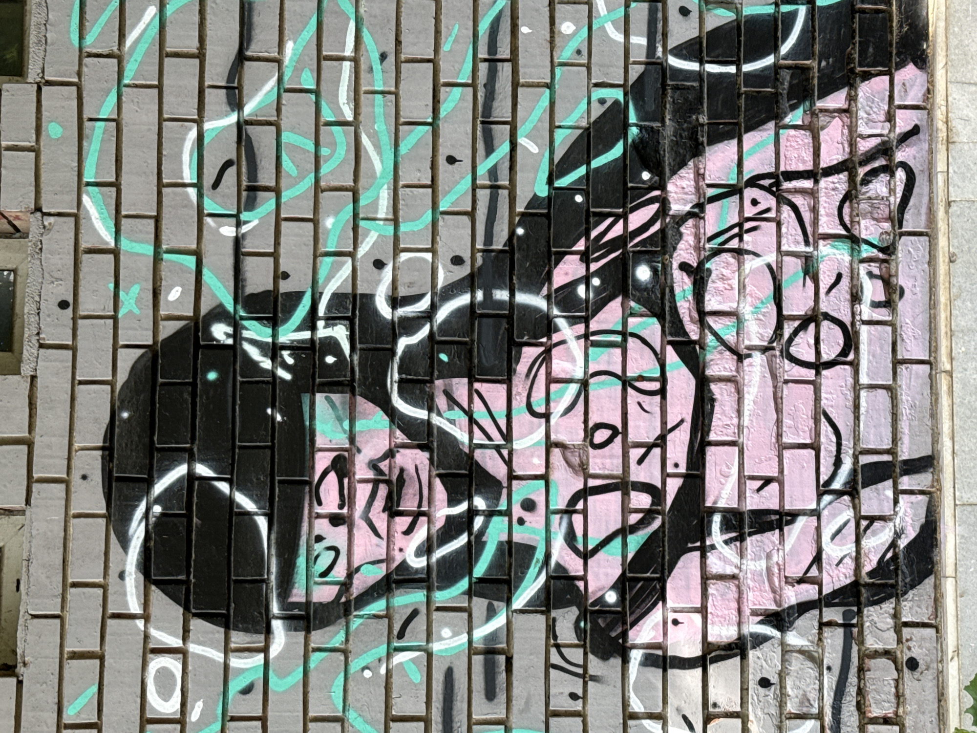 The image is a close-up, eye-level shot of graffiti on a brick wall. The graffiti depicts a stylized, pink-skinned figure with black outlines, wearing a black helmet-like head covering. The figure appears to be in a seated or hunched position, with visible hands and arms. Teal and white lines, resembling abstract swirls or energy trails, are also spray-painted across the wall, intertwining with the figure. The brick wall is constructed of light gray bricks with visible mortar lines. The lighting suggests it is daytime, with shadows indicating a light source from the upper right. The location of this graffiti in Pleven, Bulgaria, is not specified in the image.