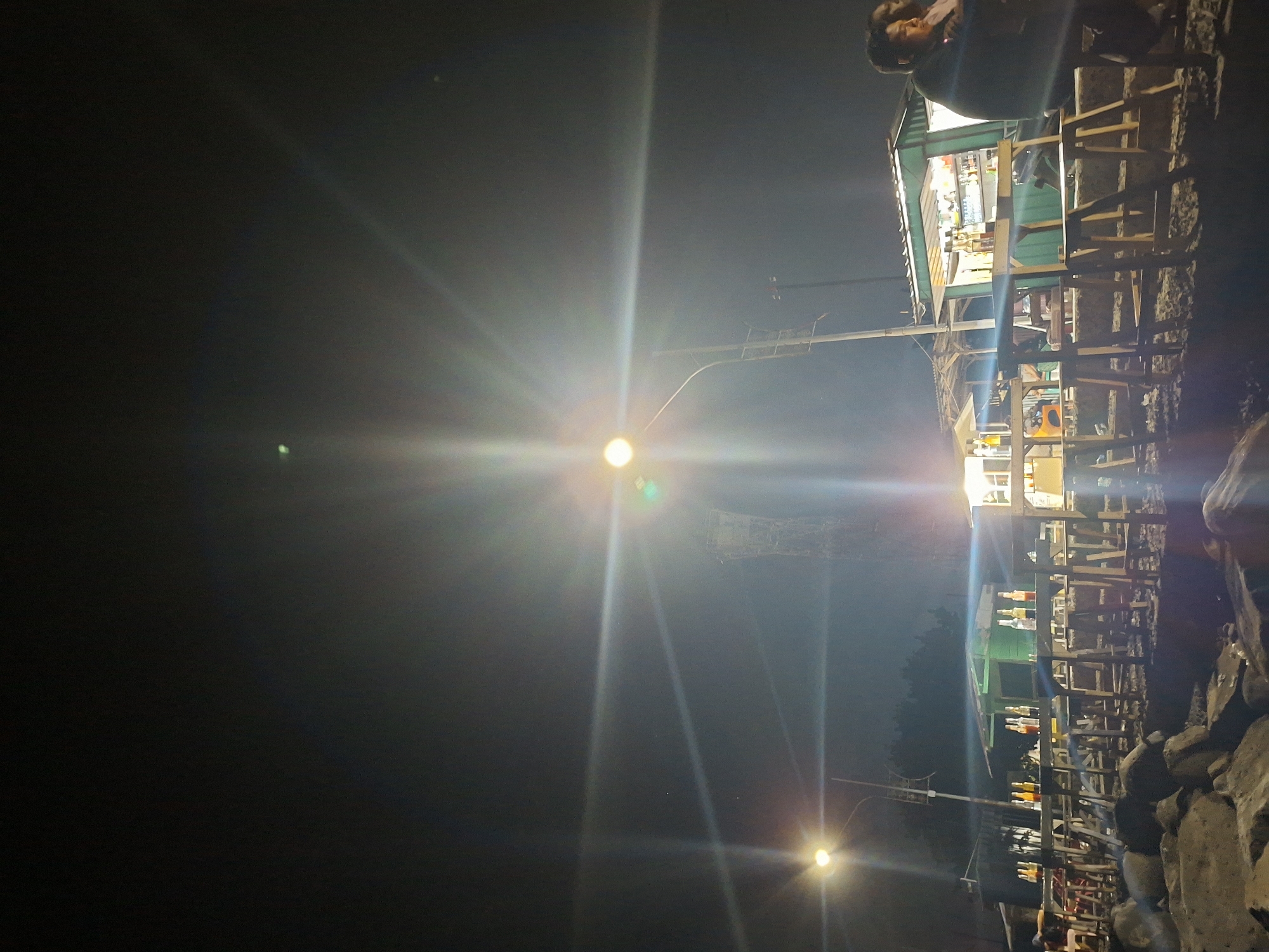This is a night scene in Lhokseumawe, Indonesia. A bright street lamp illuminates a wooden structure that appears to be a beachfront shop or restaurant. The structure has a light green roof and is elevated on stilts. Numerous shelves display bottles of liquids, likely beverages. A person is visible at the top of the frame near the structure. To the right of the building is a rocky area, suggesting a coastal location. The rest of the scene is very dark.