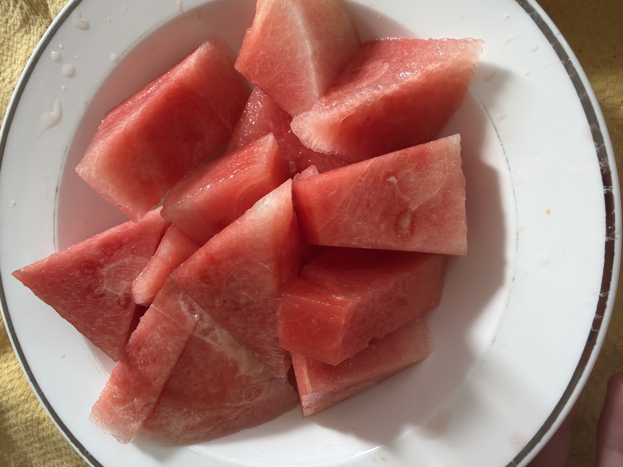 A white plate with a double-banded black and grey rim is filled with chunks of cut watermelon. The watermelon is a deep pinkish-red color, and some pieces have a visible, watery sheen. The plate is placed on a yellow textured surface. A finger is visible in the bottom right corner of the image. The image appears to be taken indoors under natural lighting. There is no indication that the image was taken in Pentre, United Kingdom.