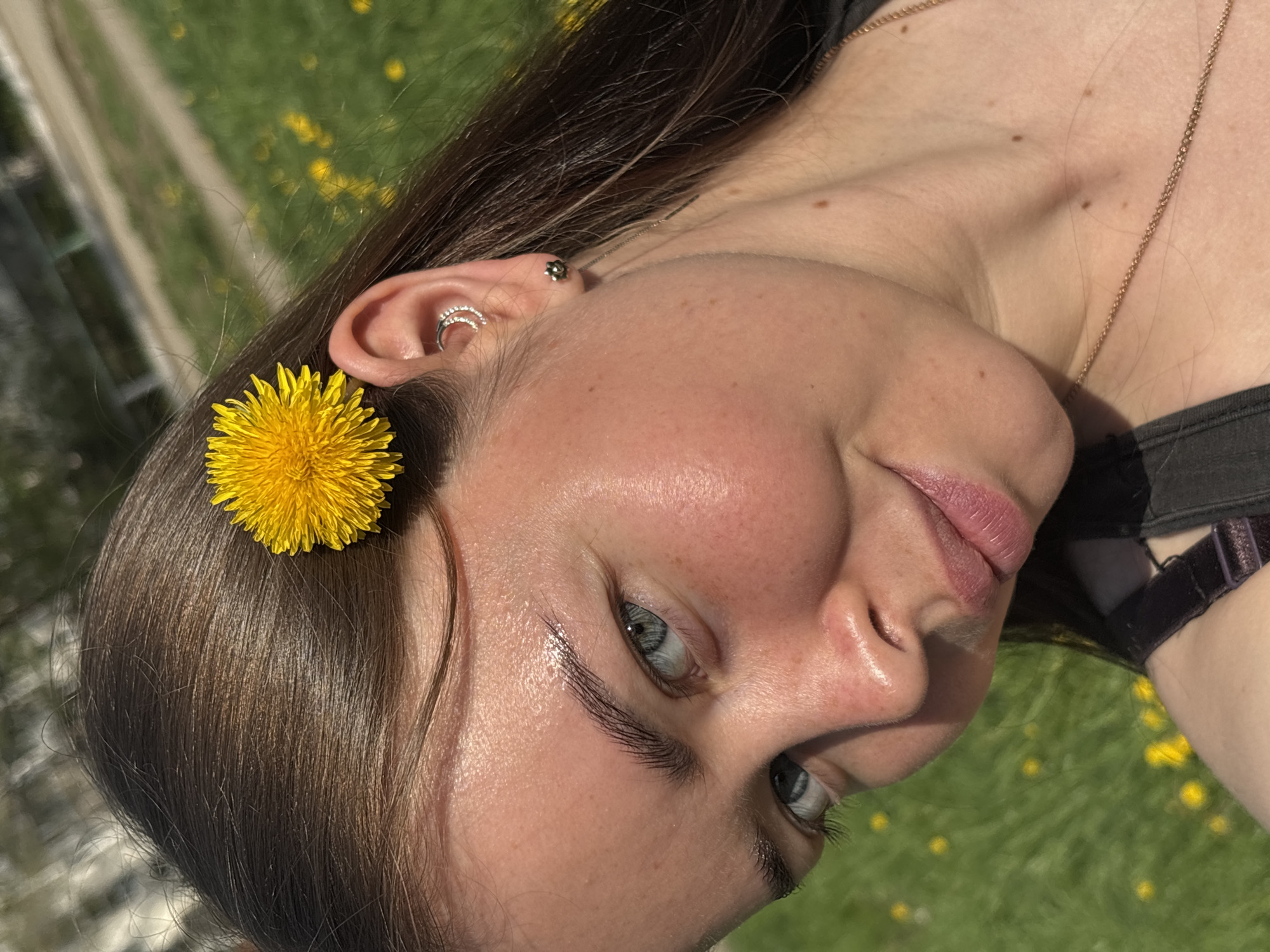 A close-up of a fair-skinned woman's face with blue eyes, turned slightly to the right, with a yellow dandelion flower tucked behind her left ear, which is adorned with a double hoop earring and a stud earring. She wears a thin gold chain necklace and a black top. The background is blurred, showing green grass dotted with small yellow flowers and a glimpse of an urban environment with a building. The location cannot be definitively identified as Izhevsk, Russia, based solely on the image.