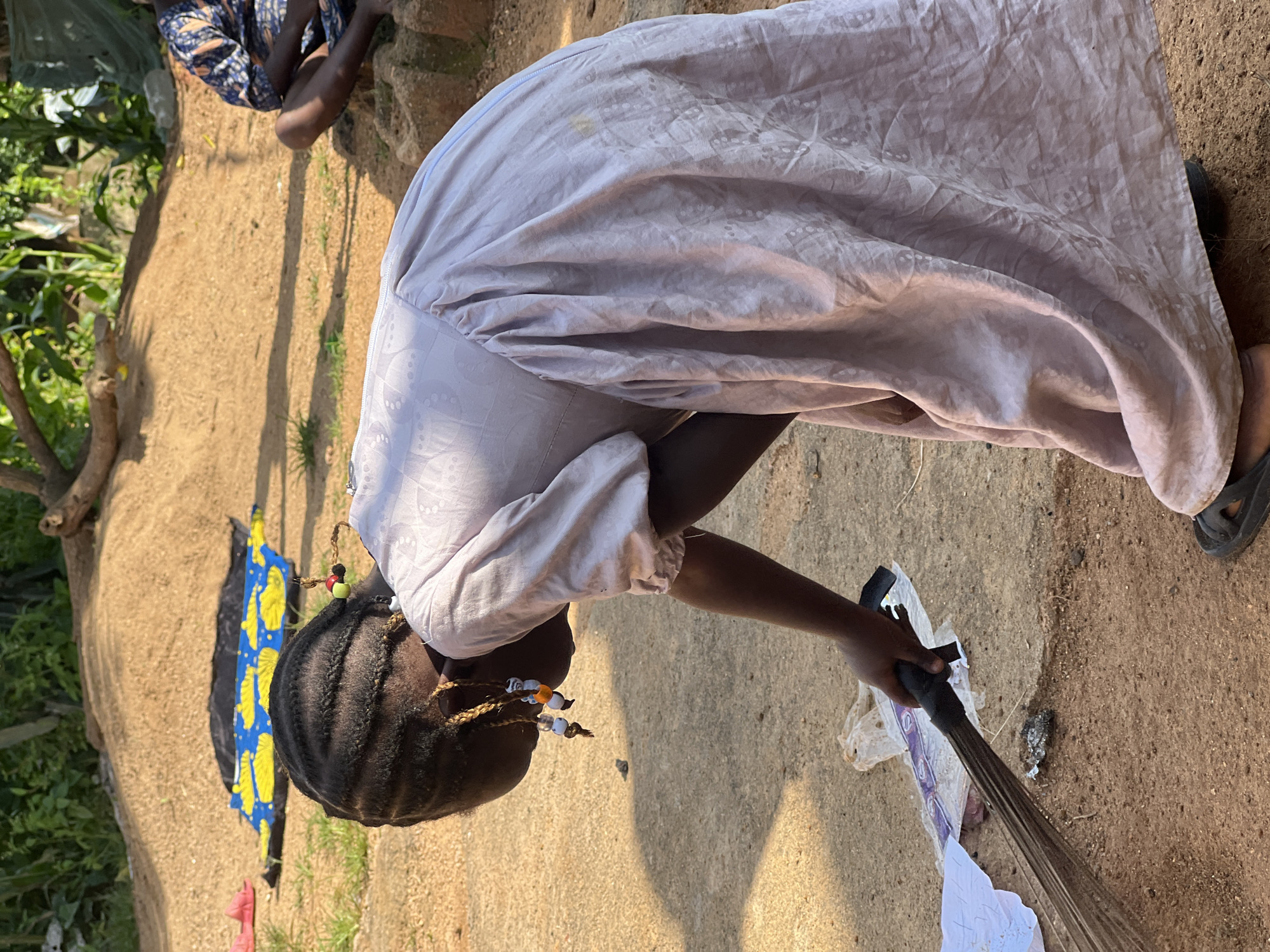 A young Black girl with braided hair adorned with beads is bending over and sweeping with a broom. She is wearing a light purple dress with a patterned texture and dark sandals. The ground is composed of dirt and some paved areas. In the background, there is a pile of sand, some green foliage, and a person sitting in the distance wearing a blue and orange patterned garment. The setting appears to be outdoors in a somewhat unkempt area, possibly a yard or a communal space.