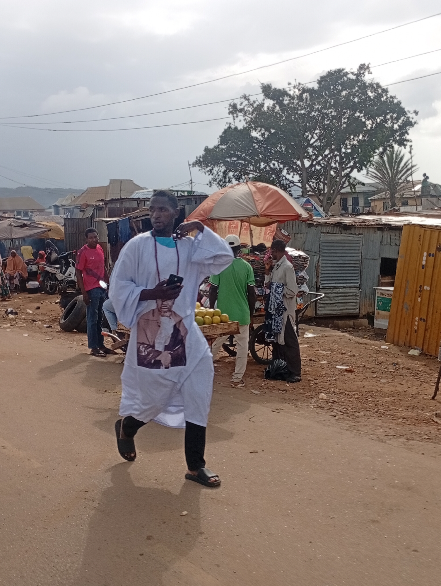 In Rafin Par, Nigeria, a man in a white robe with a portrait printed on the front walks on a dirt road. He holds a black smartphone in his right hand. Behind him, another man in a pink t-shirt stands near a motorcycle. To the right, a vendor with an umbrella sells fruit from a cart. The background shows a collection of makeshift structures with corrugated metal roofs, typical of a marketplace. The sky is overcast.