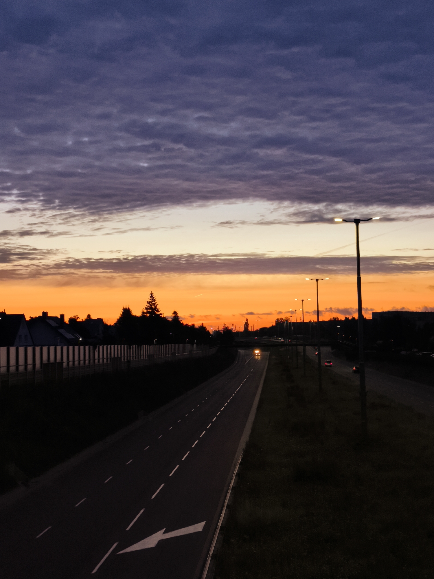 The image depicts a twilight scene on a suburban highway near Gdańsk, Poland. The sky is a dramatic blend of deep purple clouds transitioning to vibrant orange and yellow hues near the horizon, indicating either sunrise or sunset. A multi-lane asphalt road stretches into the distance, with white lane markings and a clear directional arrow visible in the foreground. Several streetlights, some illuminated, line the right side of the road, casting a faint glow. On the left, a line of houses with dark roofs is partially visible behind a tall, slatted sound barrier. Silhouetted trees, including a prominent evergreen, stand between the houses and the road. A few cars, their headlights or taillights illuminated, are present on the highway, suggesting moderate traffic. The overall atmosphere is one of transition, capturing the fading light of day or the first signs of dawn.