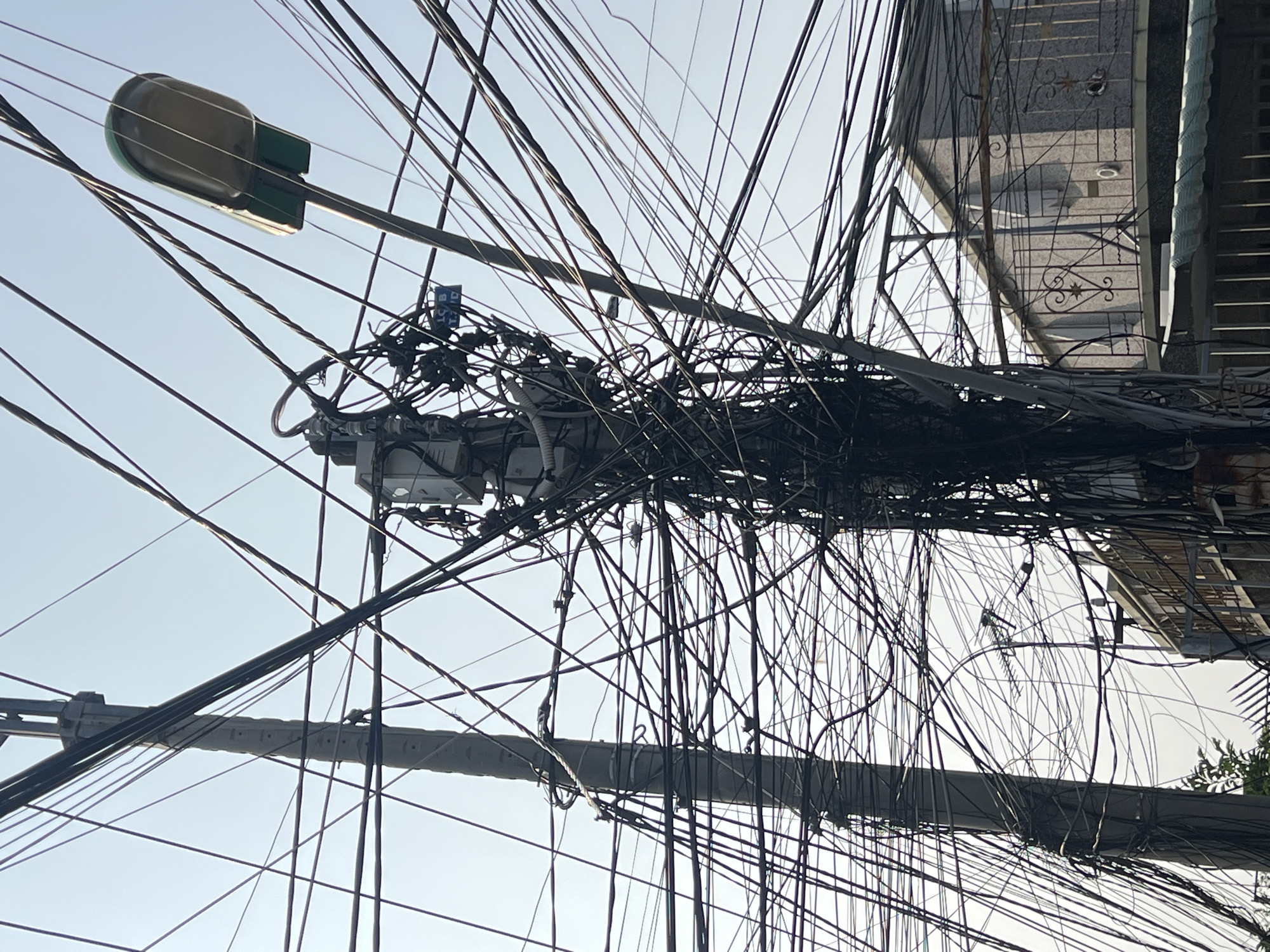 The image depicts a dense network of overhead utility wires in Bắc Nha Trang, Vietnam. The wires are tangled and disorganized, creating a chaotic visual effect against a pale sky. A street light is visible on a pole, with a cylindrical head and green accents. Adjacent to the wiring cluster is a partial view of a building with a gated balcony. The environment suggests an urban setting with a mix of infrastructure and residential structures.