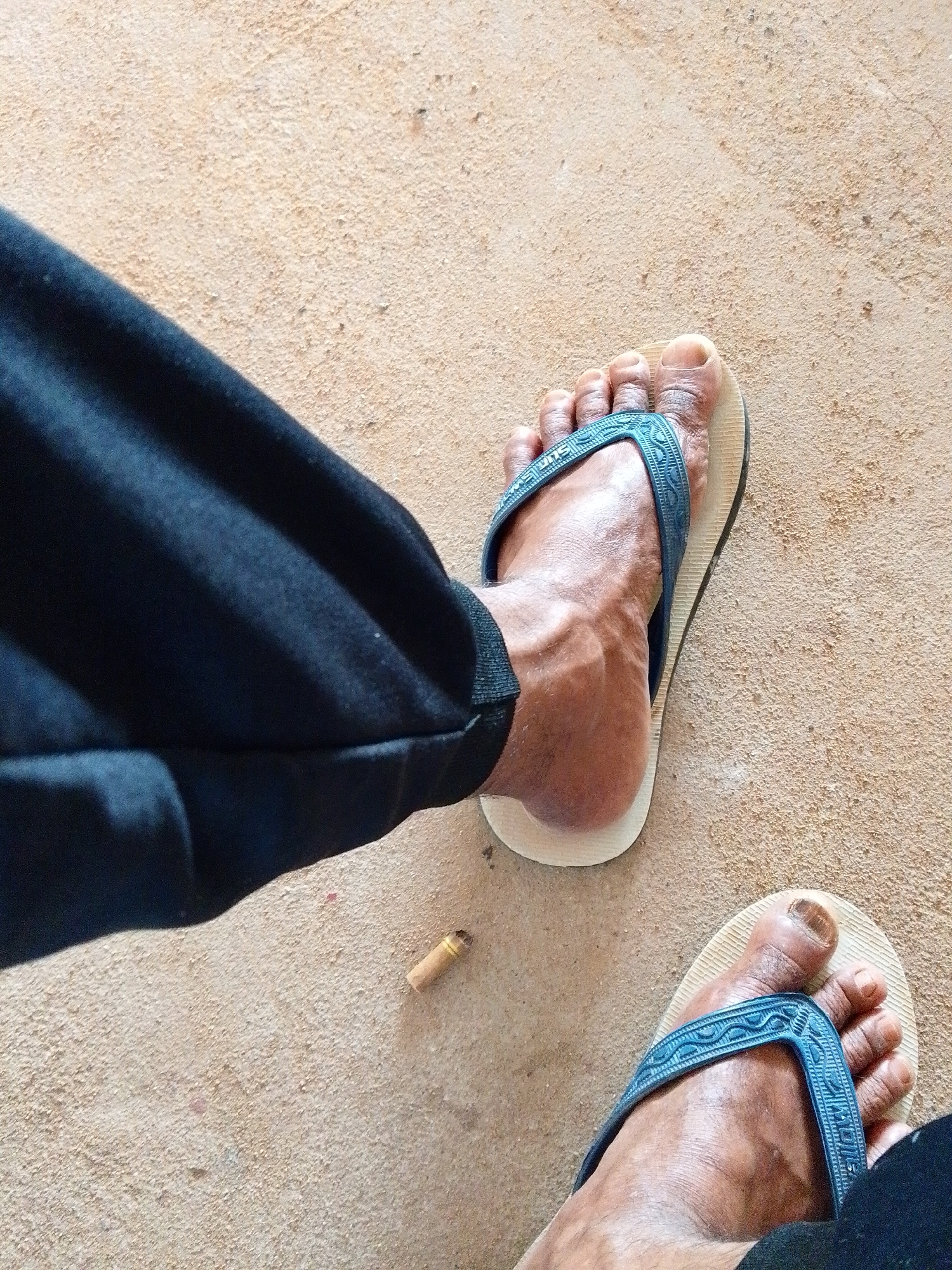 A top-down, eye-level shot shows a person's legs and feet on a neutral beige surface, possibly concrete. The person is wearing black pants that are rolled up at the ankle. Their feet are clad in off-white flip-flops with blue straps. There is a discarded cigarette butt on the ground between their feet. The setting is Bukit Ketok, Indonesia.