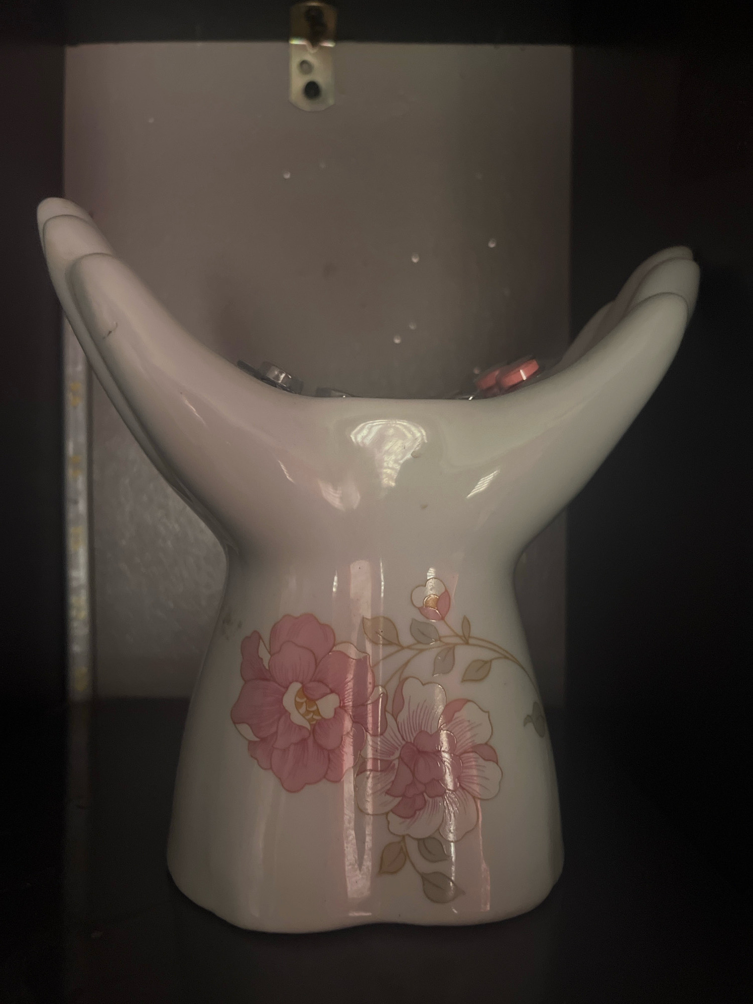 The image shows a white ceramic vase shaped like two cupped hands reaching upwards, decorated with pink floral patterns. The vase is filled with small, round objects, possibly coins or beads, some of which are red. The vase sits on a dark surface, possibly a shelf, against a neutral-colored background that appears to be inside a cabinet or on a wall. The lighting is dim, creating shadows. There is no visible indication of the city of Jos, Nigeria, or any specific urban environment.