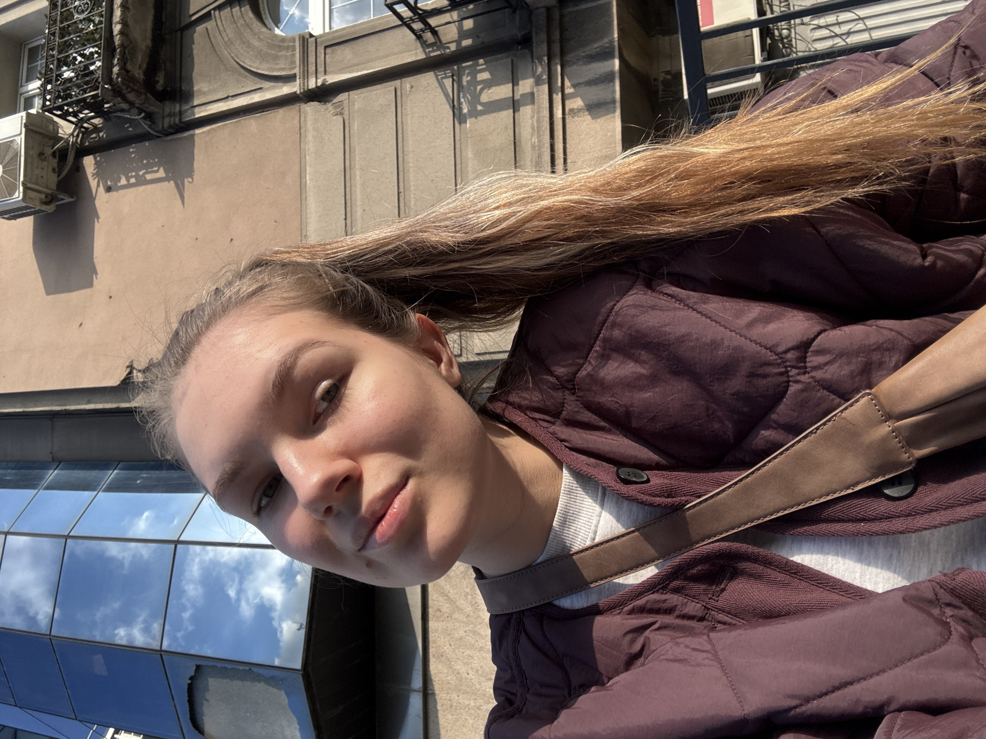 The image is an upward-angled selfie of a young woman with long, blonde hair wearing a maroon quilted jacket over a white shirt. A brown leather strap crosses her torso. In the background, buildings are visible, including a modern glass building reflecting the sky and a beige-colored building with classical architectural details like an air conditioning unit, windows, and decorative elements. The setting is likely an urban environment, possibly Belgrade, Serbia, based on the architectural style of the buildings and the urban context.