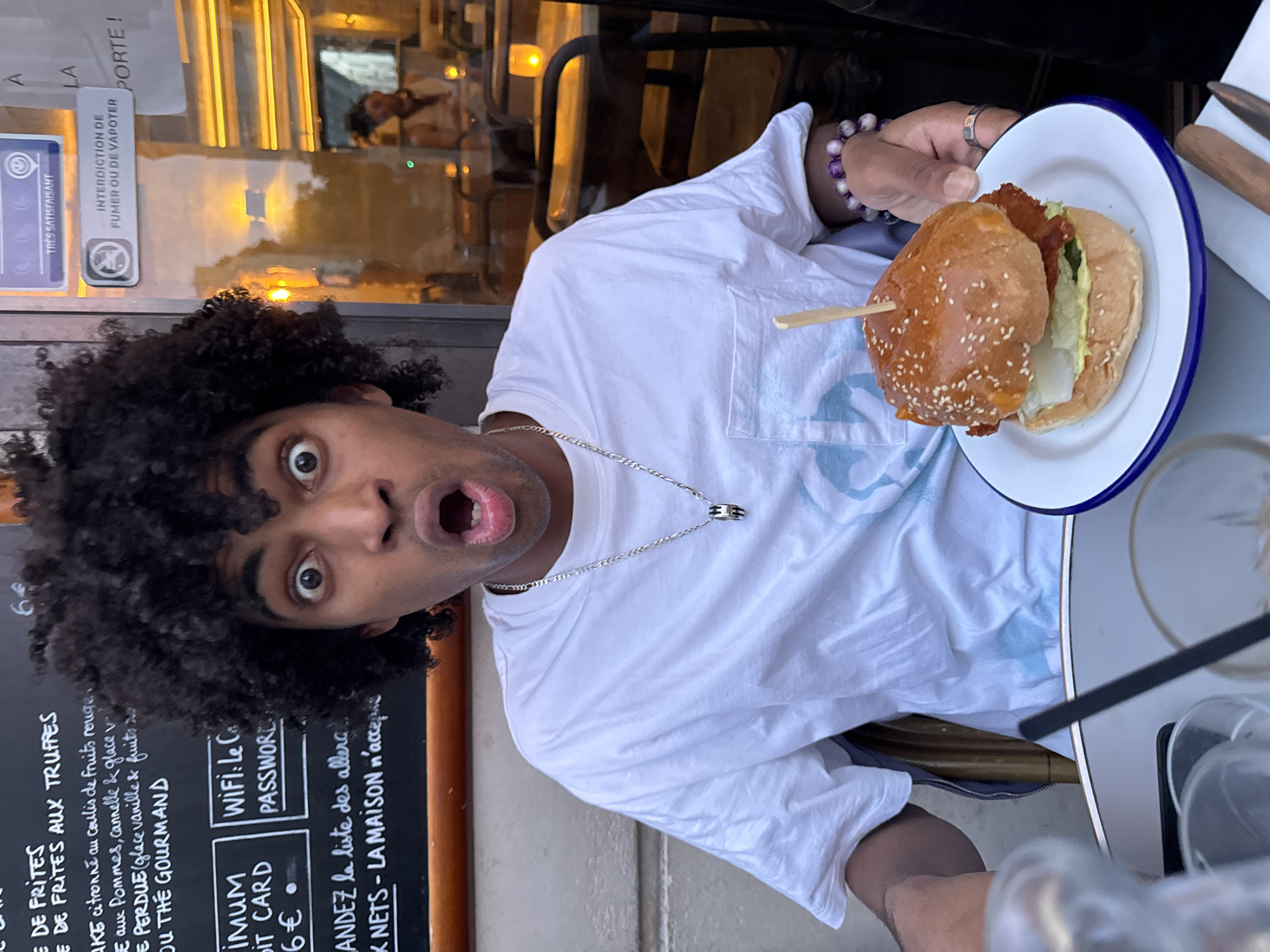 A close-up shot captures a man with curly hair and an open mouth, exhibiting a surprised expression. He's wearing a white t-shirt with a silver necklace and a bracelet with purple beads. He is holding a plate with a burger. A wooden stick pierces the burger. A sign is visible behind the man. The sign lists "DE FRITES AUX TRUFFES" and mentions "Wi-Fi" and "PASSWORD." The lower part of the sign says, "minimum credit card 6 euro." Another sign prohibits smoking or vaping. The photo was taken outdoors in Paris, France.