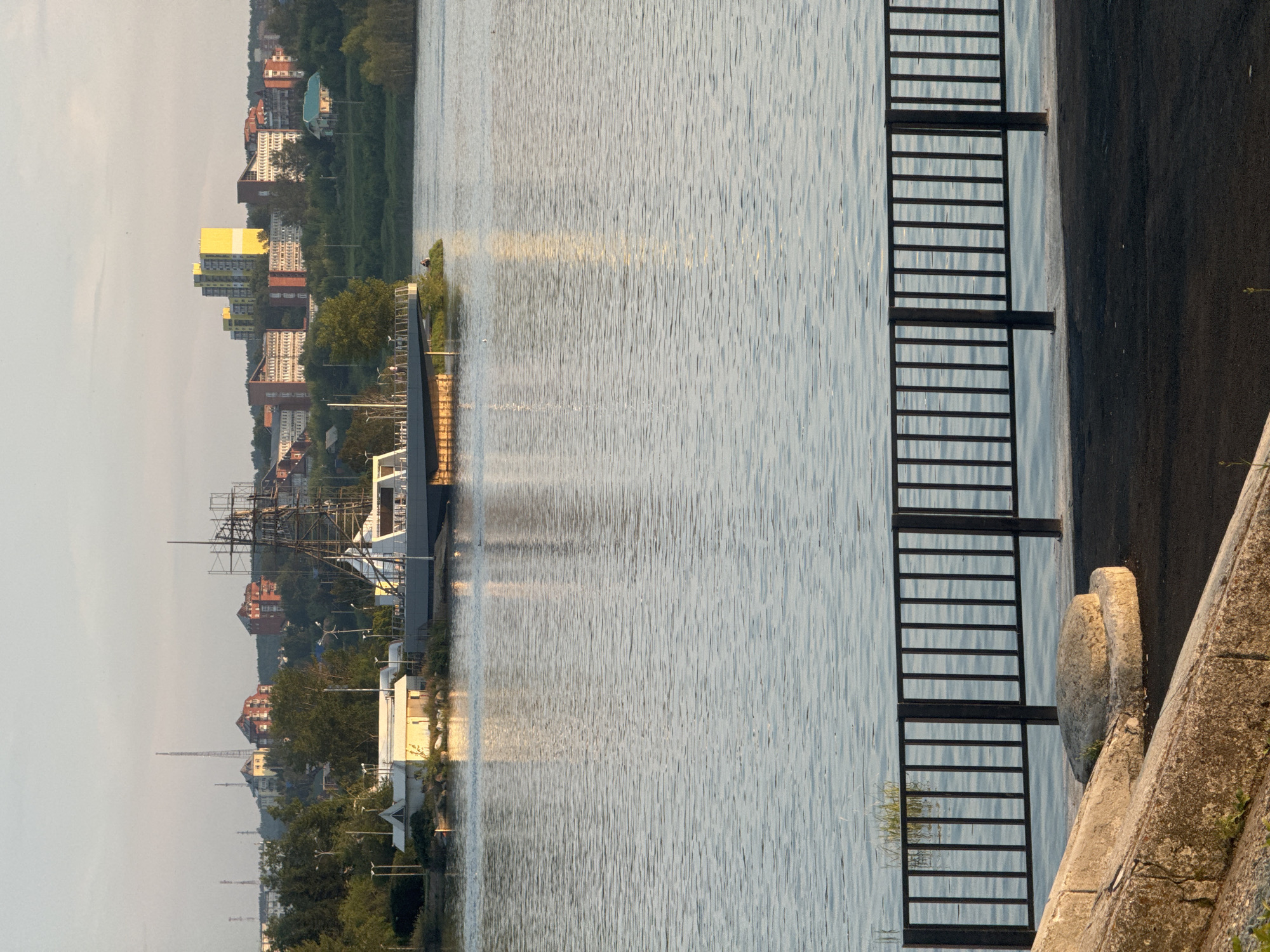 The image captures a serene view of the Angara River in Irkutsk, Russia. In the foreground, a black metal railing separates a paved walkway from the rippling water. Beyond the railing, the river's surface reflects the soft, hazy light of what appears to be early morning or late evening.
On the far bank of the river, a city skyline is visible. Dominating the water's edge is a peculiar structure that resembles a partially constructed ship or a historical replica, complete with scaffolding. Adjacent to this, a small, white building with a yellow accent is situated on a rocky embankment dotted with greenery.
Further inland, a cluster of buildings forms the city's backdrop. These range from smaller, older brick structures to more modern, multi-story apartment buildings, including a striking yellow and blue high-rise that stands out against the softer hues of the sky. The landscape is generally characterized by gentle hills covered in trees, indicating a city integrated with its natural surroundings. The overall atmosphere is tranquil and peaceful.