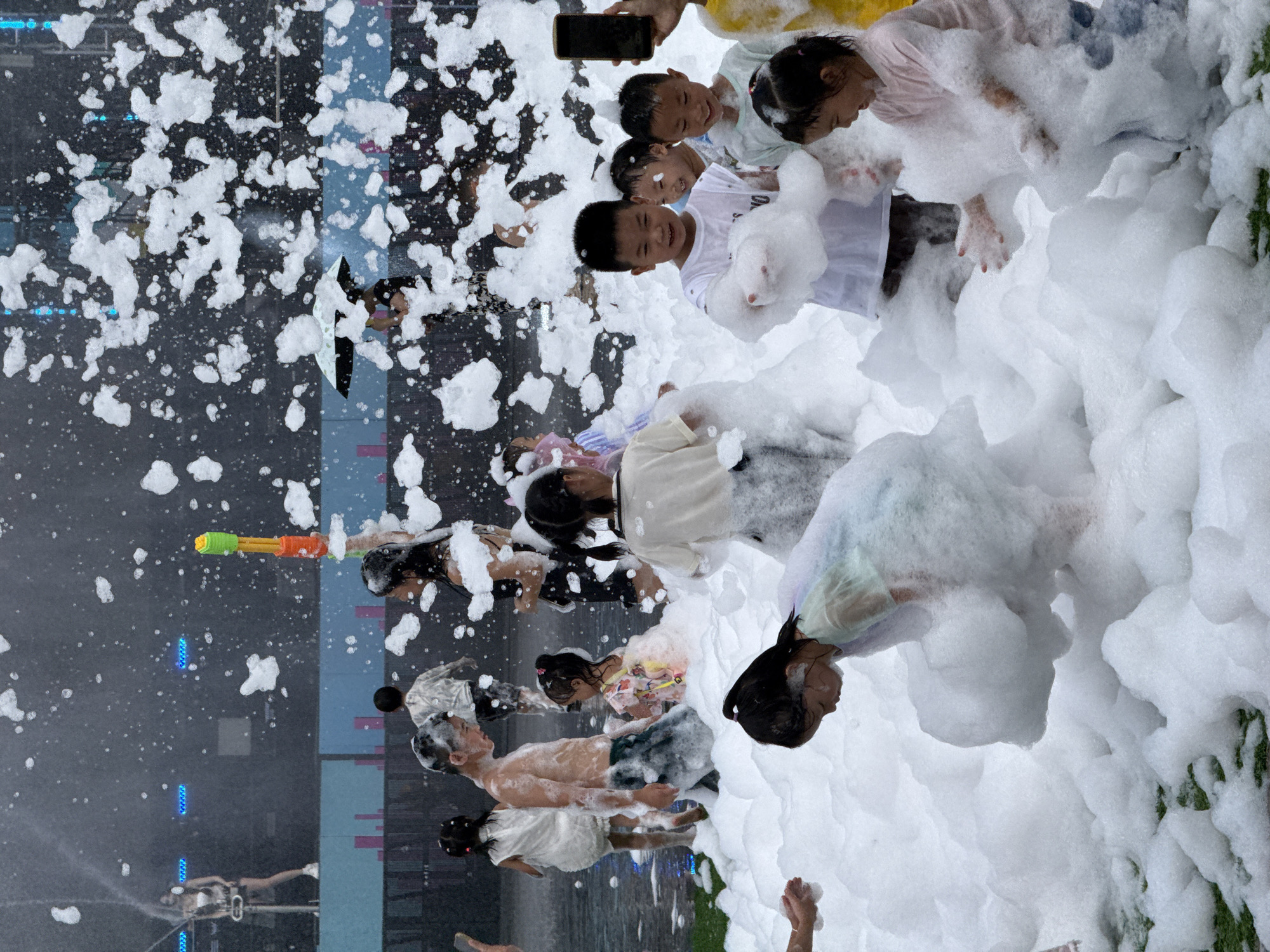 In Nanshan District, China, a group of children are joyfully playing in a large foam party. The scene is filled with white, fluffy foam, creating a playful and chaotic atmosphere. Several children are visible, many of whom are young boys and girls, immersed in the foam. Some are smiling and interacting with each other, while others are looking around or playing with the foam. In the background, there are glimpses of adults, a stage with lights, and some sort of structure or backdrop, suggesting this event is taking place outdoors in a public or entertainment area. The lighting appears to be natural, perhaps during the daytime or early evening. The overall impression is one of fun and excitement, with the foam serving as the central element of the activity.