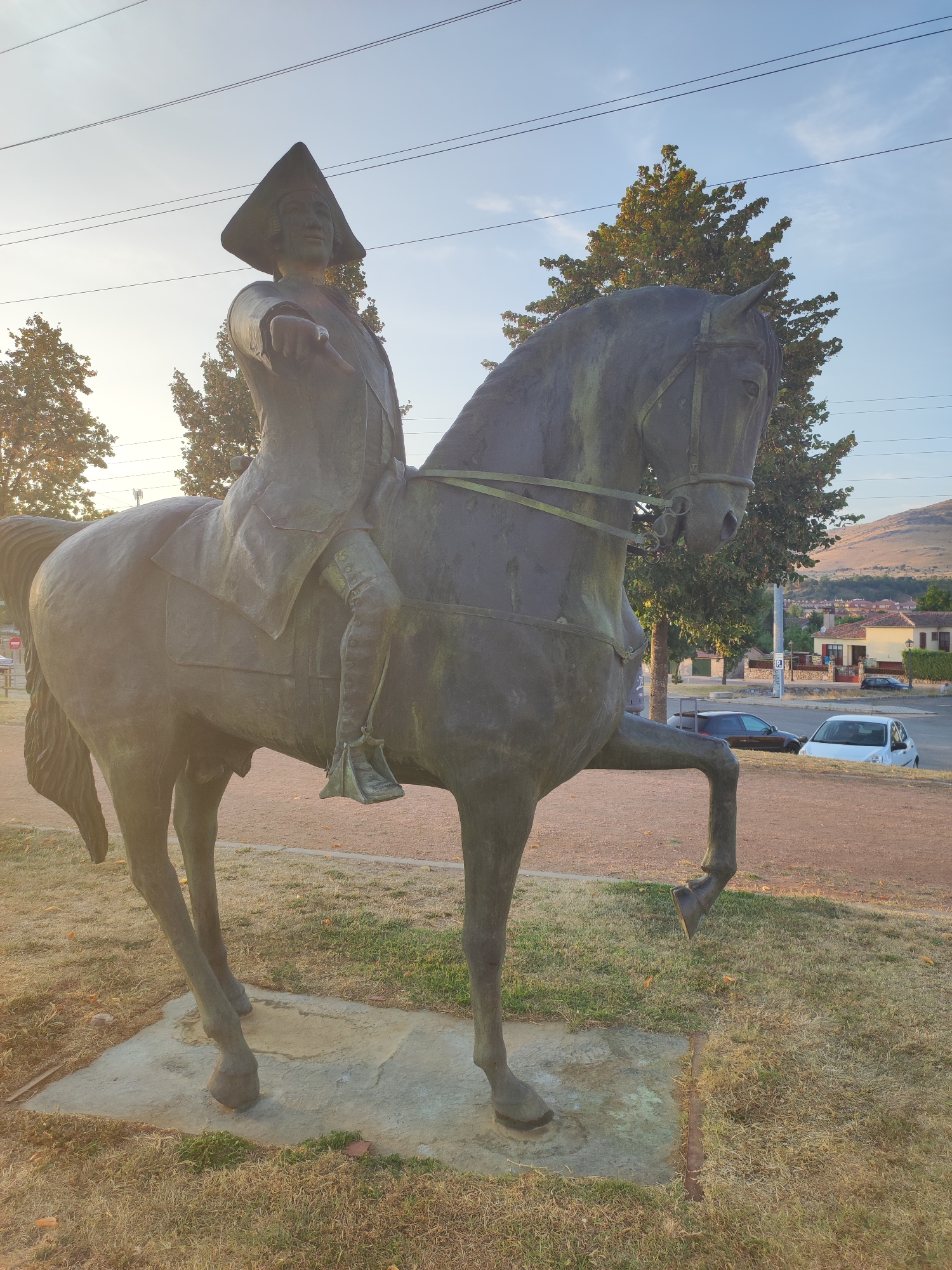 A bronze equestrian statue of a man wearing a tricorn hat and a long coat is positioned on a concrete base in a grassy area. The man's right arm is extended forward, pointing. The horse is depicted with its left foreleg raised. In the background, there are trees, buildings with red tile roofs, a street with cars, and a hillside under a clear sky. The statue is located in the city of San Ildefonso, Spain.