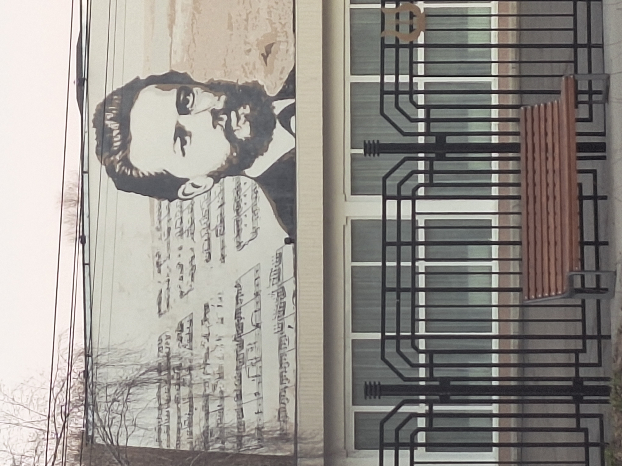 This image presents a mural of a bearded man, presumably a historical figure, positioned next to a building facade. The building features a series of windows with an ornate black metal grating and a wooden bench attached. The background contains a few leafless trees and power lines, suggesting it might be in a more urban setting, specifically Unknown, Unknown. The mural appears to be painted on a wall of a building, with musical notes depicted below the man's portrait. The composition is somewhat obscured due to the angle and the presence of the building structure.