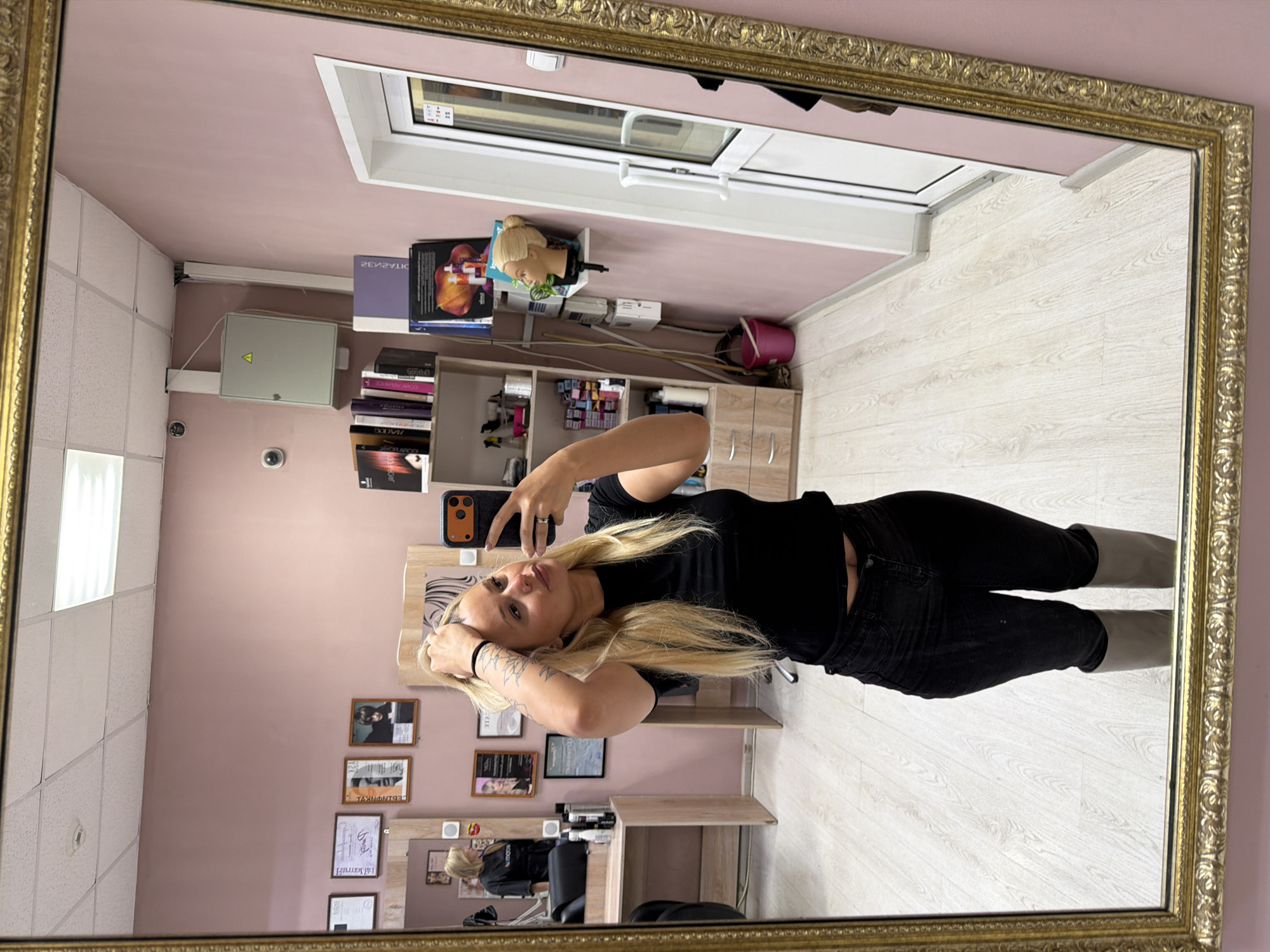 A young woman with blonde hair and tattoos is reflected in a large, ornate, gold-framed mirror. She is wearing a black t-shirt and black jeans, holding a smartphone in her hand and making a hand gesture. The reflection shows the interior of what appears to be a salon or beauty parlor. The walls are light pink, and there's a tiled ceiling on one side. In the background, there is a bookshelf with various items, including a mannequin head, along with documents hung on the wall. A window is seen above the bookshelf. The floor is light-colored wood. A salon chair and table are visible in the lower part of the reflection. This photograph was most likely taken in Sarapul, Russia.