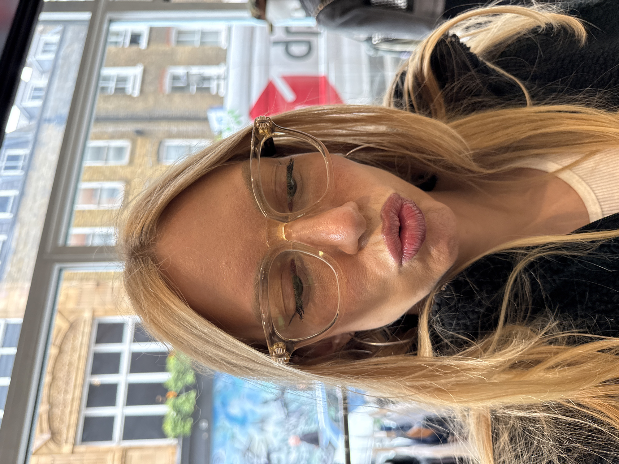 A woman with long blonde hair and clear-framed glasses is captured in a close-up, possibly a selfie, inside a building. Her head is tilted, and her lips are pursed in a slight pout. Her eyes, visible behind the clear lenses of her glasses, appear to be closed or looking downwards. She is wearing a dark, possibly black, fluffy or textured jacket over a light-colored (white or off-white) top, the collar of which is visible.
The setting appears to be indoors, next to a large window with white frames. Through the window, an urban environment is visible, suggesting a city location. The background shows multi-story buildings with numerous windows. One building has a light brown or beige facade with white-framed windows, while another to the left is a lighter, possibly greyish-blue color. The background is slightly out of focus, drawing attention to the woman.
The natural, bright lighting suggests it is daytime. In the blurry background, visible through the window or as a reflection, there is an inverted red triangular shape with some indistinct white text, possibly part of a sign like "dp" or similar, indicating a business. The architecture in the background is consistent with an urban area like London, United Kingdom. The image conveys a posed, perhaps contemplative or stylish, moment.
