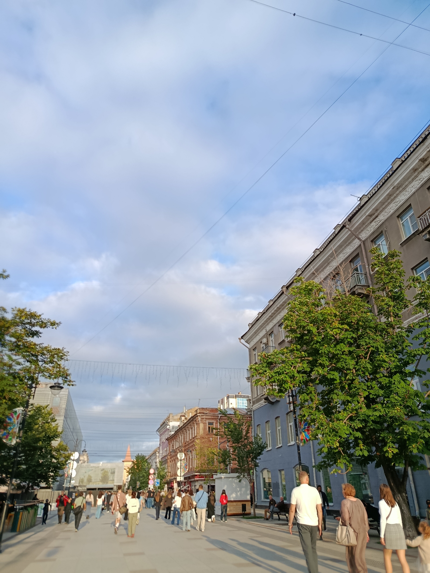 This image depicts a pedestrian street in Saratov, Russia. A diverse group of people are walking along the street, some are standing and talking, and others are sitting on benches. The street is lined with buildings of various architectural styles and heights, and there are trees and streetlights visible. The sky is partly cloudy, with the sun casting long shadows across the pavement. In the background, a tall, conical tower with a red roof stands out against the skyline. The scene suggests a lively urban environment, possibly during a warm day, with people enjoying the public space.