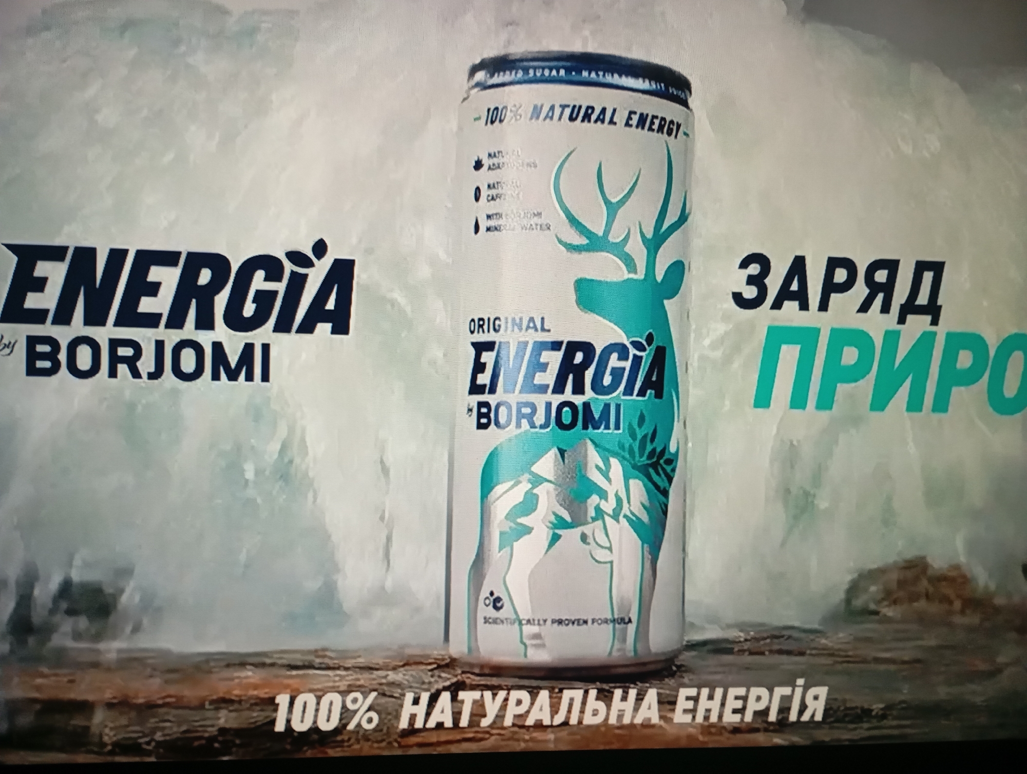 In an advertisement for "Energia by Borjomi," a can of the drink is prominently displayed. The can features a teal deer graphic and text indicating "100% NATURAL ENERGY." To the left of the can, the brand name "ENERGIA by BORJOMI" is written in large, dark blue letters. To the right, the Ukrainian text "ЗАРЯД ПРИРО" (likely "ЗАРЯД ПРИРОДИ" or "Charge of Nature") is visible in large, teal letters. The background is a stylized representation of a natural landscape, possibly with a geyser or waterfall, rendered in shades of white and light blue, suggesting a cold or natural environment. The bottom of the image shows a rustic, wooden surface with more Ukrainian text reading "100% НАТУРАЛЬНА ЕНЕРГІЯ" (100% Natural Energy). The scene appears to be a studio-shot advertisement. There are no people or identifiable urban elements present.