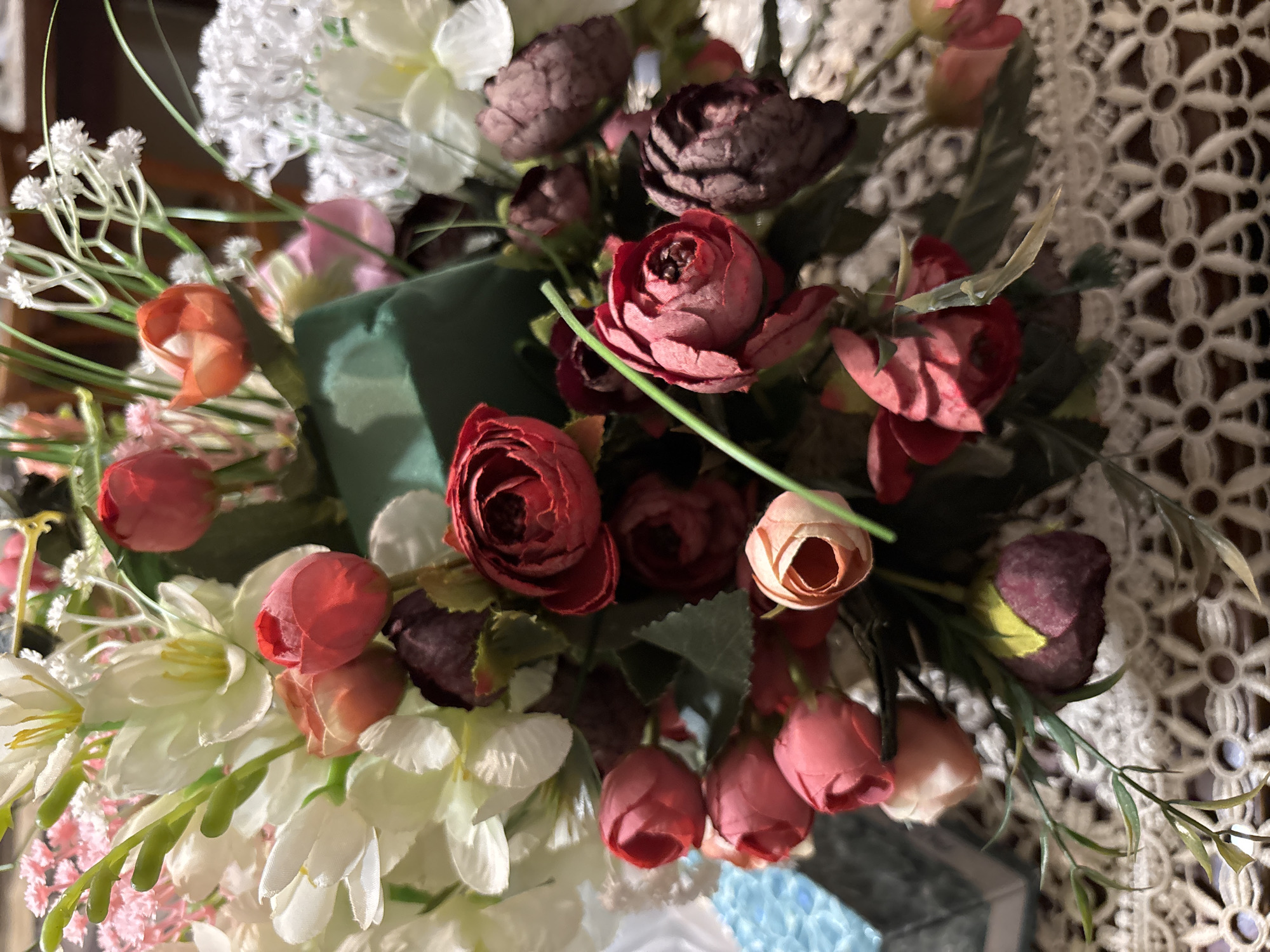 This close-up image showcases a vibrant artificial floral arrangement, displaying a mix of various flower types and colors. The dominant elements are deep red and burgundy artificial roses, some in full bloom revealing their detailed, layered petals, and others as tightly closed buds. These are complemented by lighter pink and peach-colored flower buds scattered throughout the bouquet.
Interspersed among the roses are clusters of delicate white flowers, resembling baby's breath, and larger white blossoms that appear to be artificial lilies or daffodils, some featuring prominent yellow stamens. Green foliage, including leaves and slender stems, provides a natural contrast to the colorful blooms. A distinct dark green element, possibly part of the arrangement's base or wrapping, is visible within the bouquet.
In the background, a white, intricately patterned lace tablecloth adds a decorative layer to the setting, featuring a repeating floral or geometric design. Towards the bottom of the frame, glimpses of a light blue object with a textured or patterned surface can be seen, hinting at additional decorative items on the surface where the bouquet is placed. The lighting suggests an indoor setting, creating soft shadows that add depth to the artificial flowers. The image captures a static display, highlighting the aesthetic appeal of the floral arrangement. No text is visible.