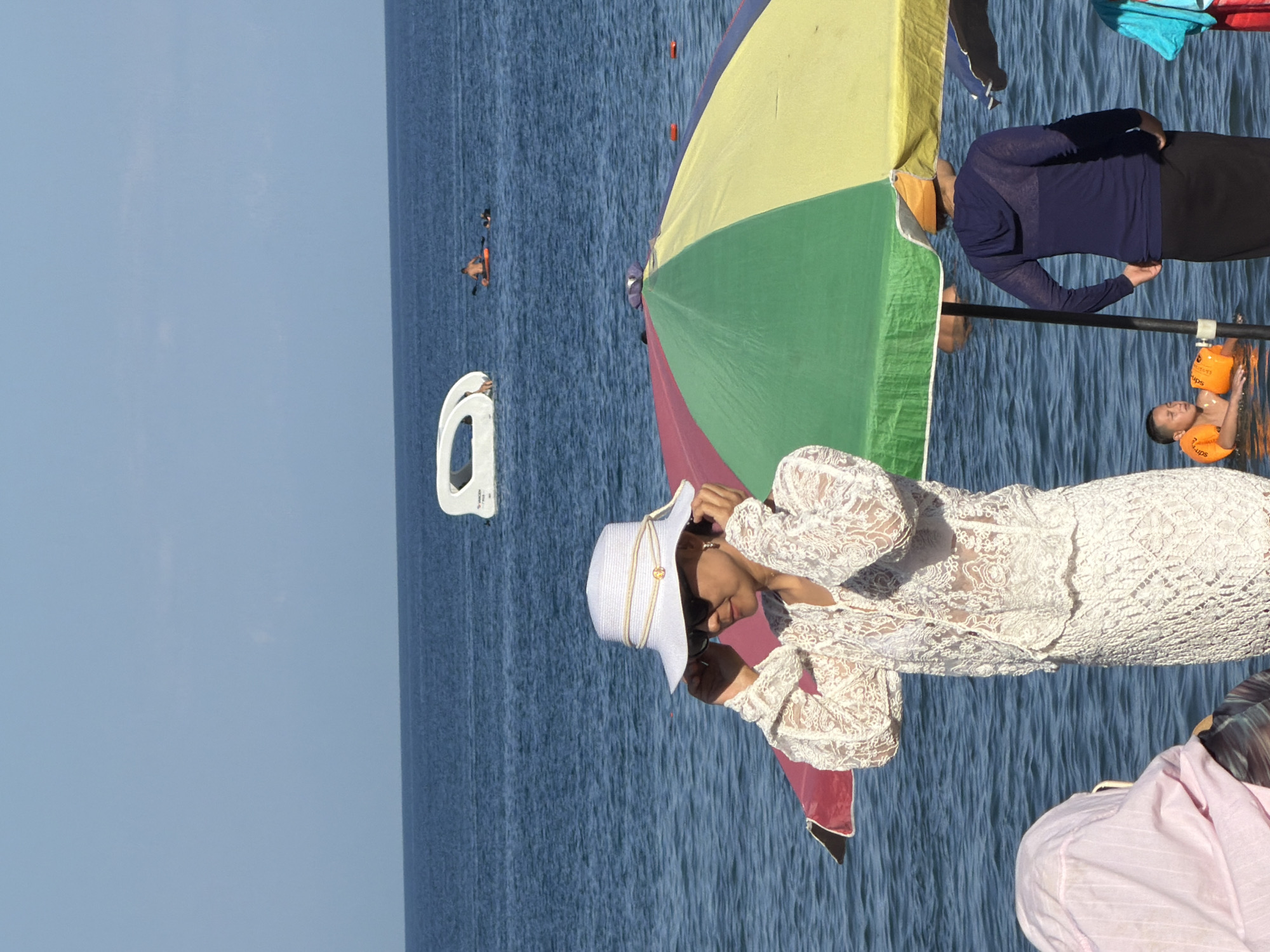 The image depicts a sunny day at a beach. In the foreground, a woman wearing a white lace dress and a white hat adjusts her sunglasses. She is standing near a colorful beach umbrella, parts of which are visible, segmented into red, green, and yellow. Behind her, the blue ocean stretches out under a clear sky. In the middle ground, a white, boat-like structure is partially submerged in the water. Further in the distance, a person is seen paddling a small boat. In the lower right of the image, another person wearing a dark blue hooded top and black skirt stands near the umbrella. To the left, a person wearing a pink head covering is partially visible. A young boy wearing orange flotation devices is in the water, looking towards the right.