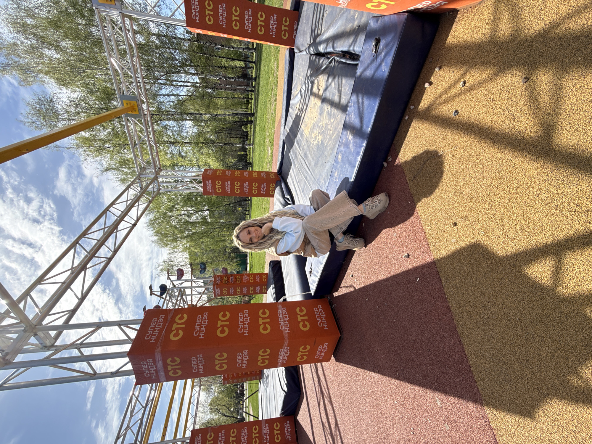 Here is a detailed description of the image:
The image shows a young woman sitting on a blue and gray platform in what appears to be an outdoor obstacle course. She has blonde hair and is wearing a light blue top and beige pants. She's smiling and looking towards the camera.
The setting includes a metal frame structure, possibly part of the obstacle course. Orange rectangular blocks with "CTC СУПЕР НИНДЗЯ" written on them are attached to the structure. Behind the course are trees and a blue sky with clouds. The ground is covered with a combination of brown and red textured material, resembling rubber flooring often found in playgrounds or sports areas.
The scene is located in the city of Шебунино, Russia.