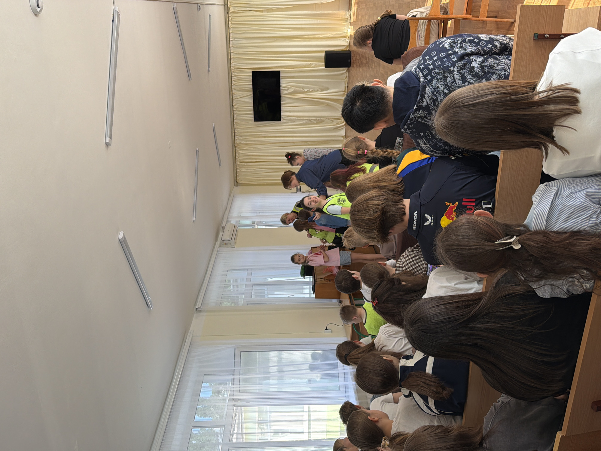Location: Classroom in Unknown, Unknown.
Scene: A classroom setting with a group of students sitting in rows of desks.
People: Several students are present, of varying ages. Some are wearing yellow vests.
Objects: Desks, chairs, windows, fluorescent lights on the ceiling, a TV screen on the wall, and curtains.
Actions: The students are seated, possibly observing something in front of them.
Interactions: The students are oriented towards a common point of focus.
Environment: The classroom appears to be well-lit.