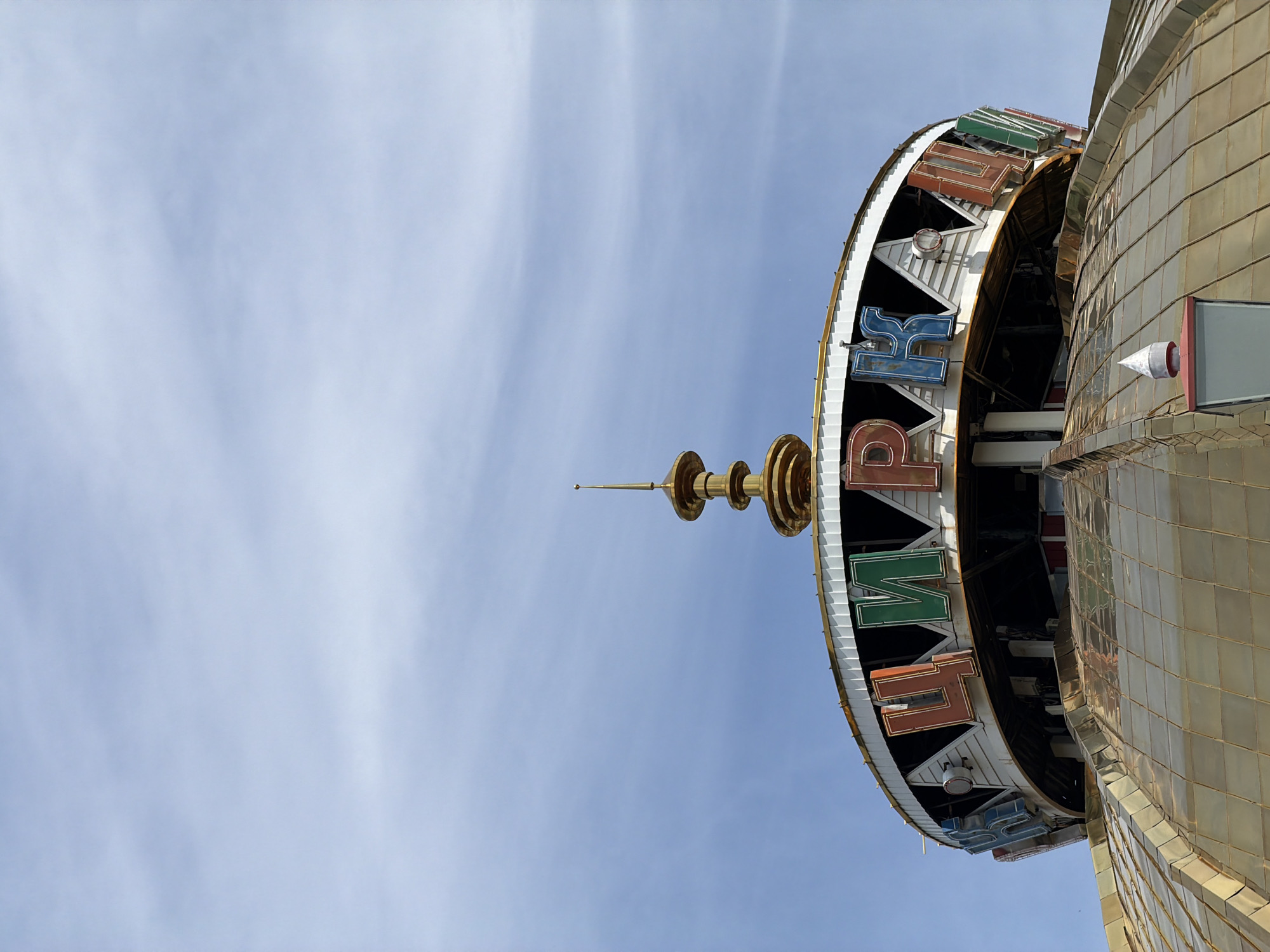 The image shows the exterior of the Khabarovsk State Circus building in Khabarovsk, Russia. The building features a domed, golden roof and a circular upper structure with colorful neon letters spelling out "ЦИРК" (Circus in Russian). A golden spire is visible at the top of the building. The sky is blue with wispy clouds. There are no people or animals visible in the image.