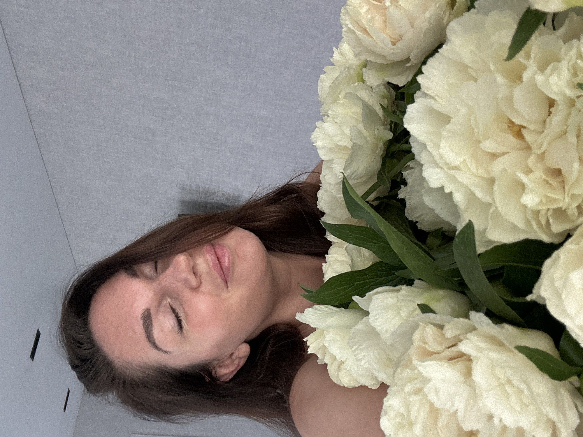 A young woman with brown hair lies next to a bouquet of white peonies. She has her eyes closed and a slight smile. The background appears to be a light gray or white wall. There is no evidence in the photo to confirm the location as Chaikovsky, Russia.